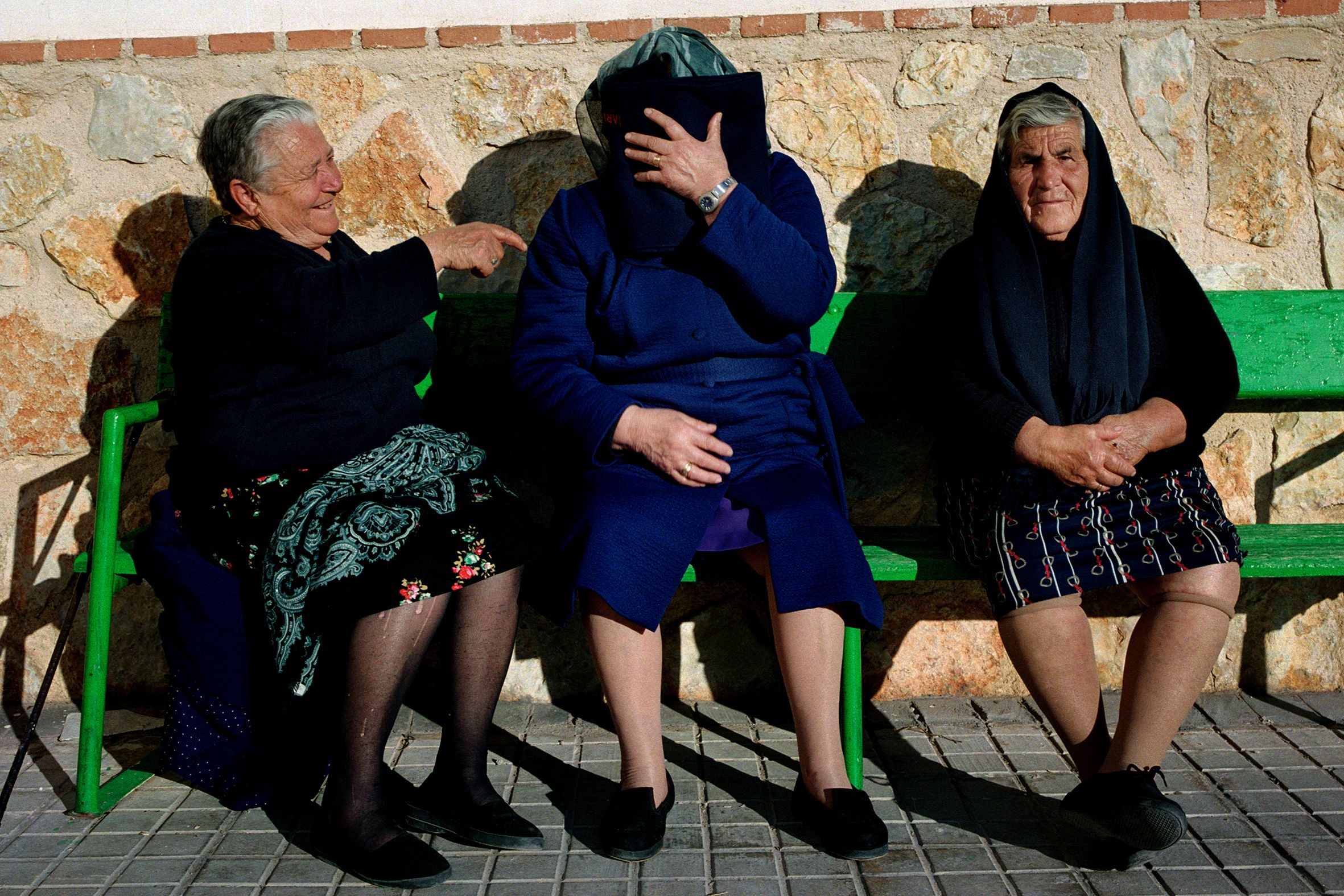 Three elderly women seated on a bench. One laughs, one covers her face with a book, the other sits with hands folded.