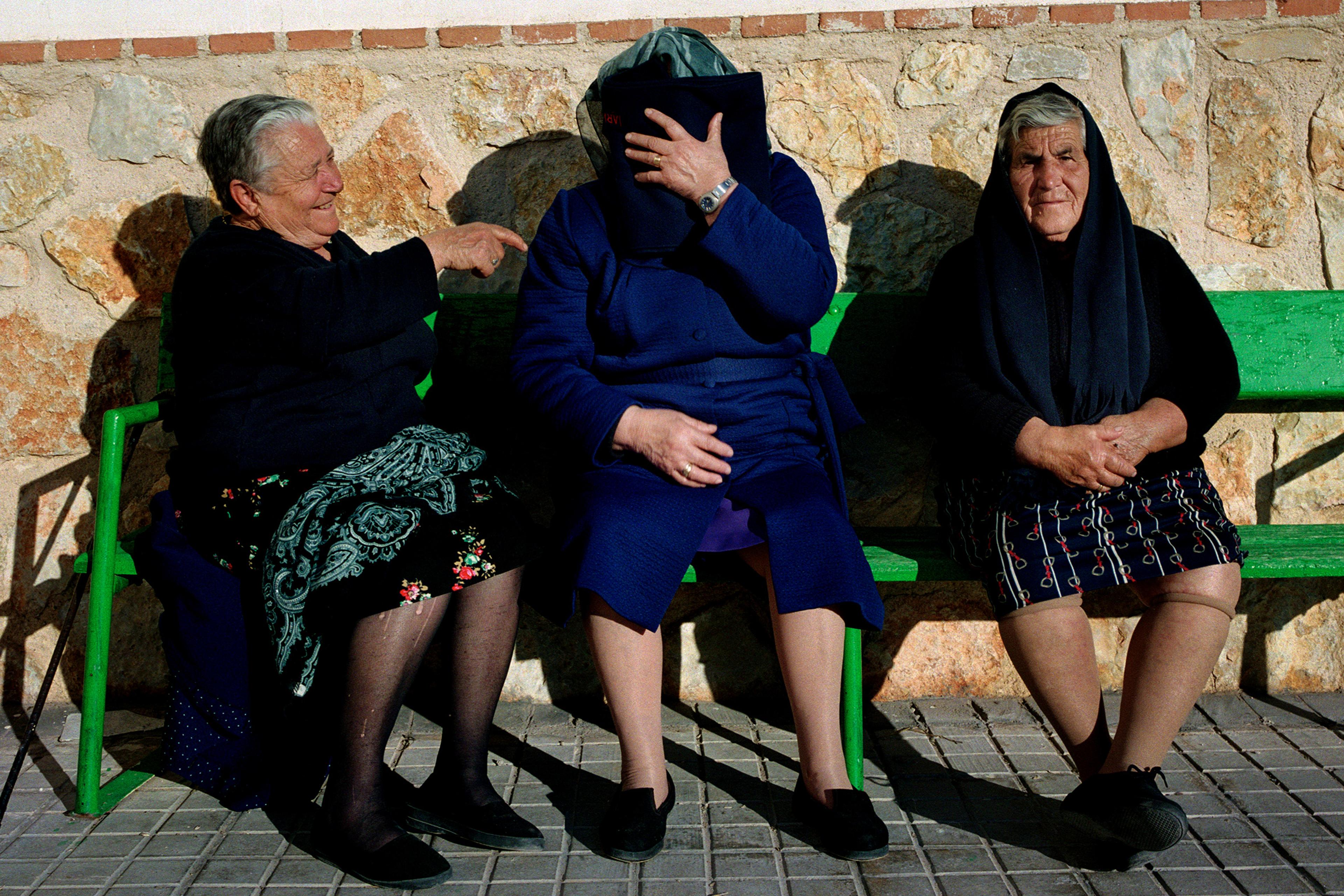 Three elderly women seated on a bench. One laughs, one covers her face with a book, the other sits with hands folded.