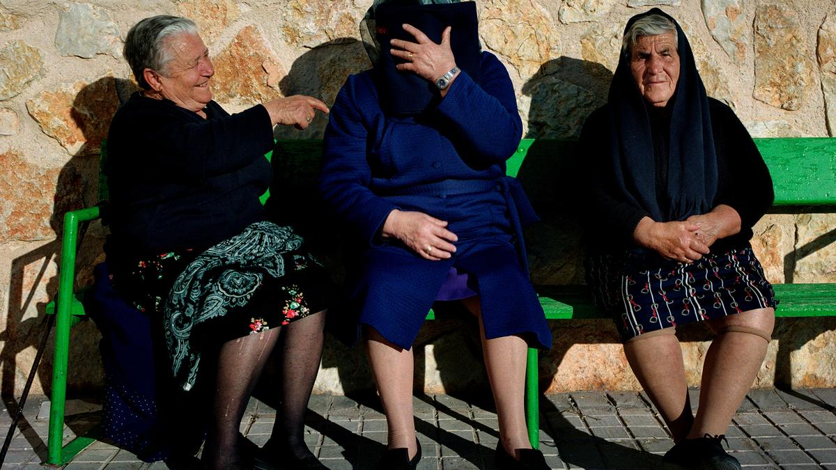 Three elderly women seated on a bench. One laughs, one covers her face with a book, the other sits with hands folded.
