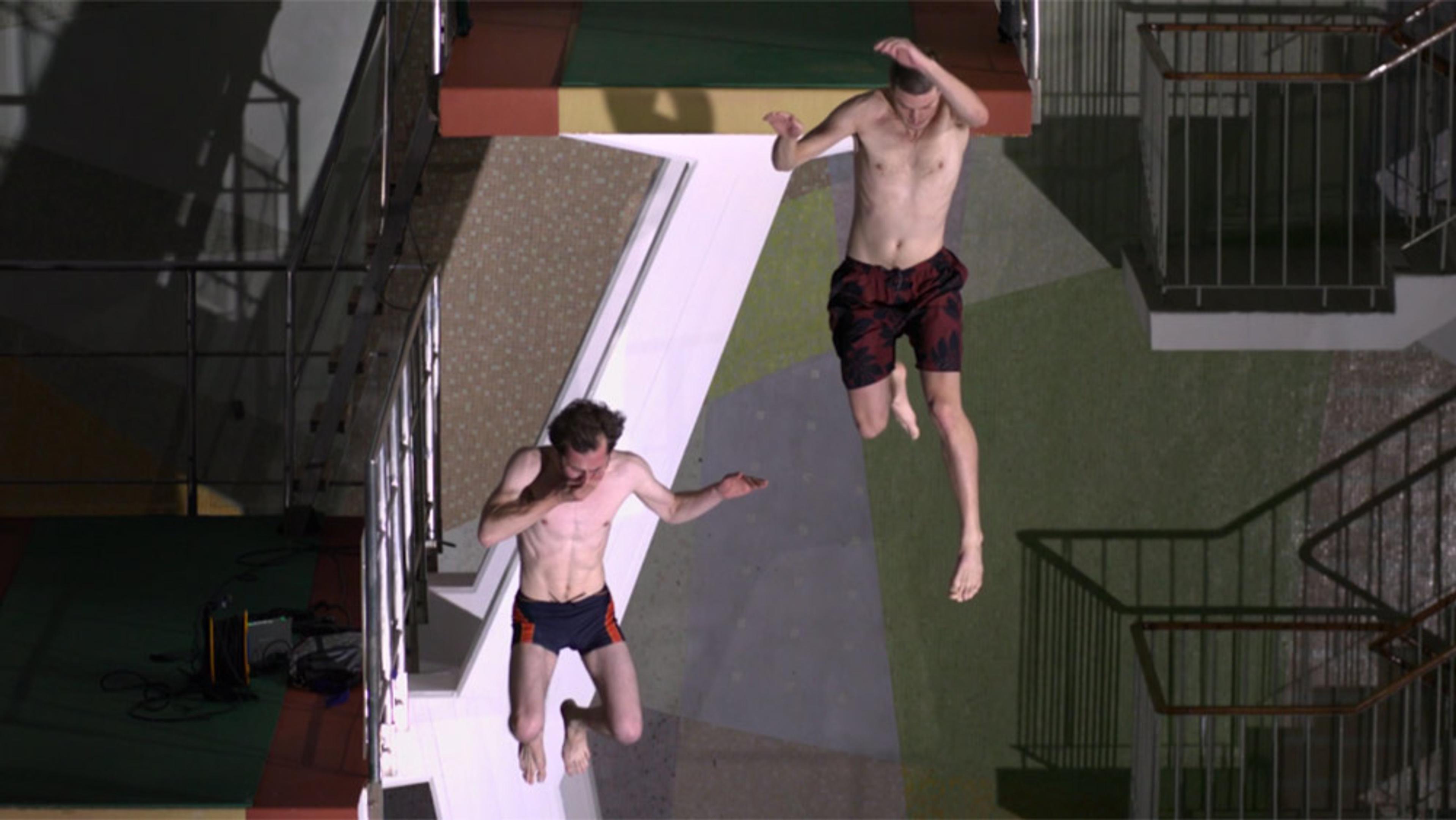 Two shirtless men in swim shorts jumping off a platform inside a building, captured mid-air with railings in the background.