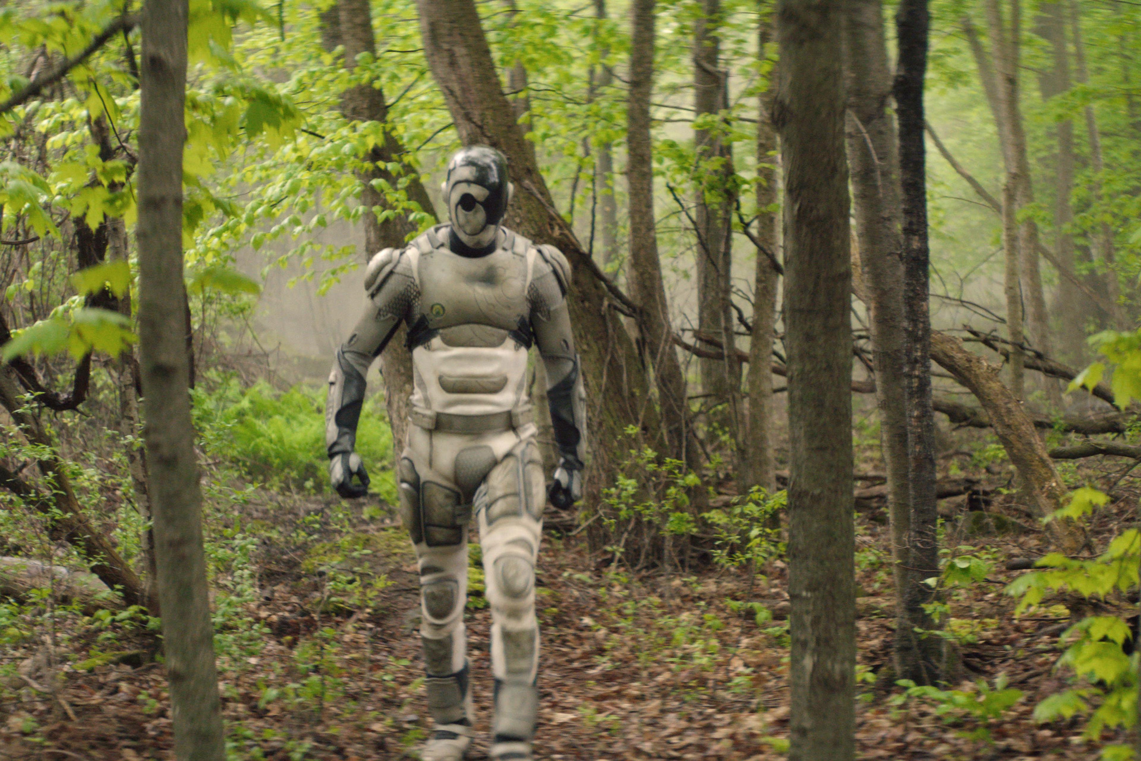 A person wearing a futuristic robotic suit walking through a lush green forest.