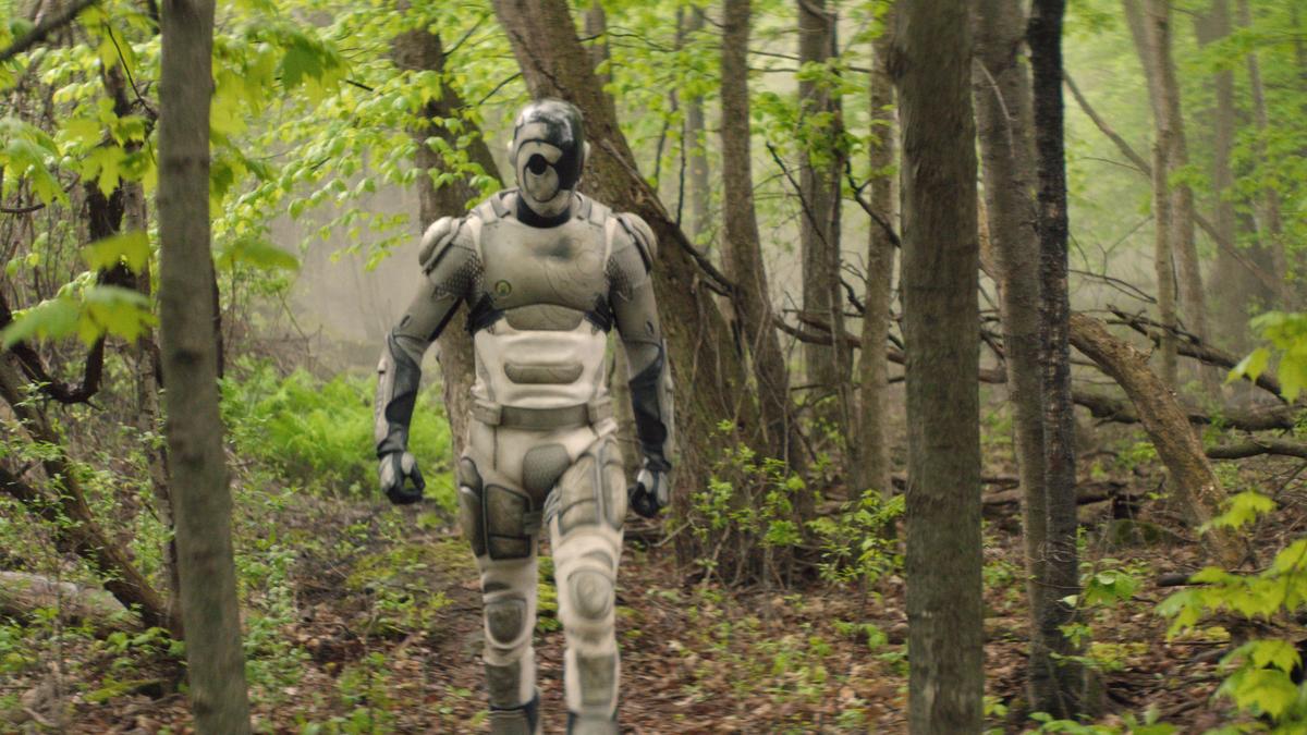 A person wearing a futuristic robotic suit walking through a lush green forest.