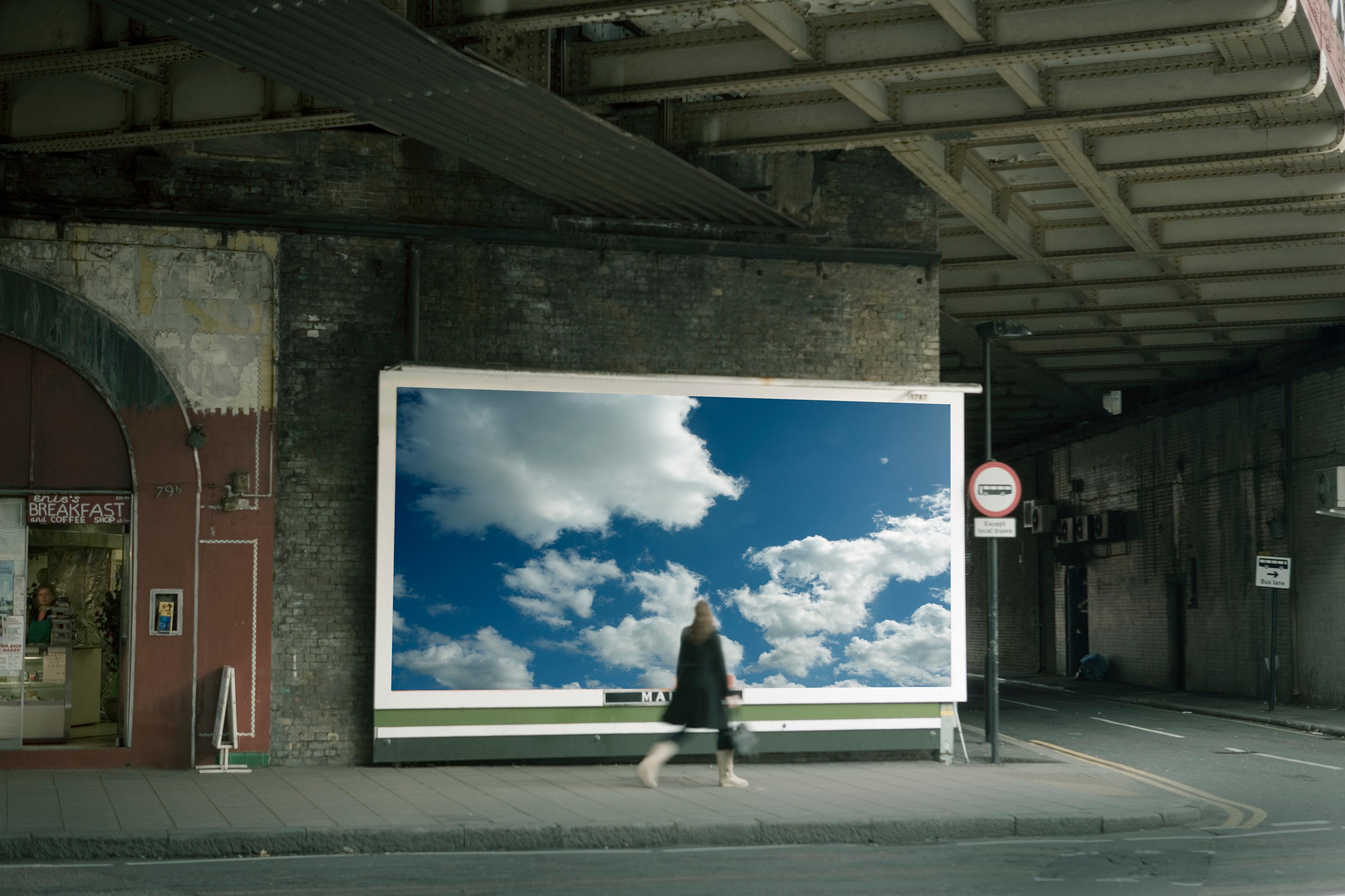 Photo of a street scene with a large billboard showing cloudy sky a woman walking by a café and an underpass roof above.