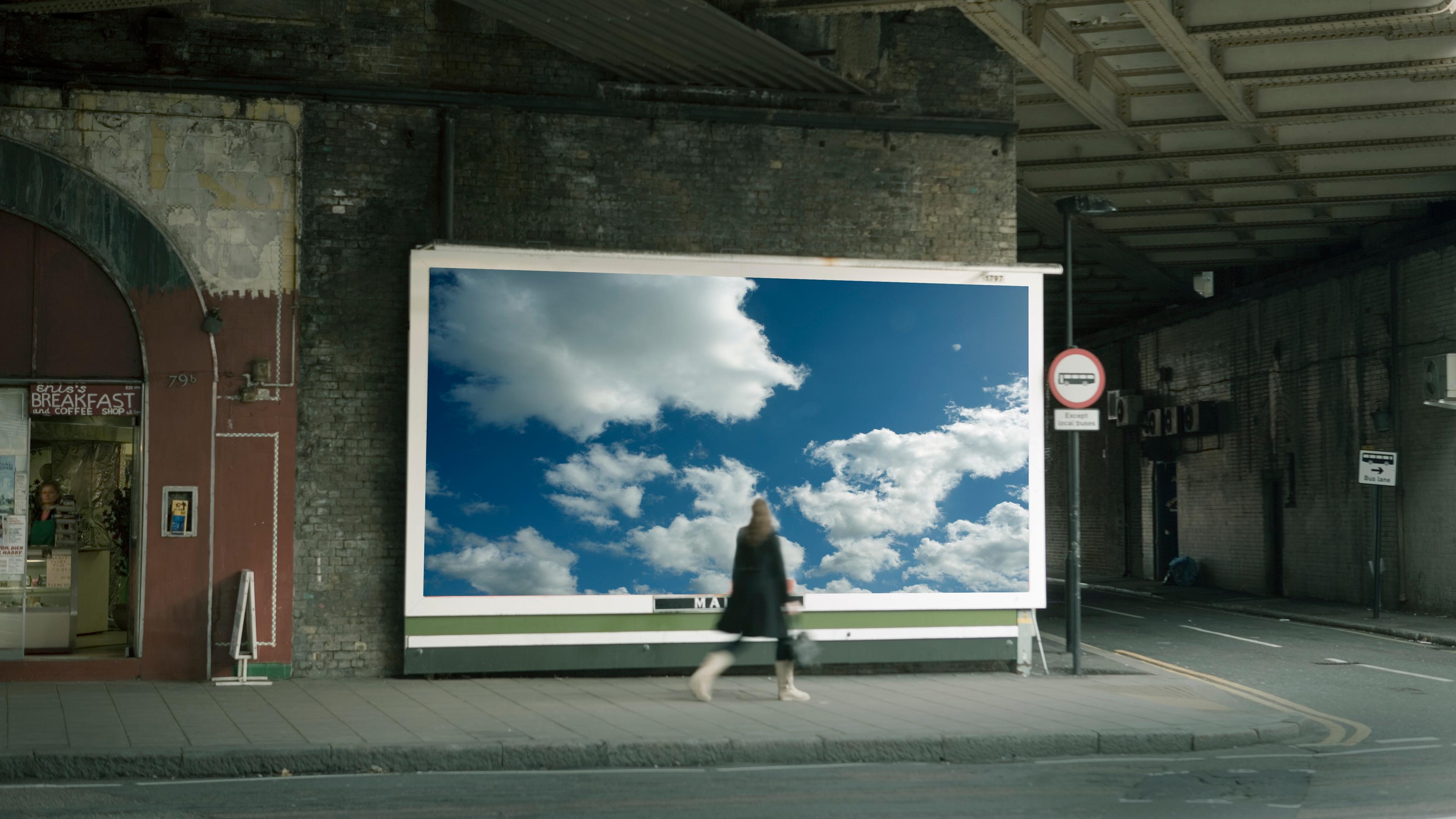 Photo of a street scene with a large billboard showing cloudy sky a woman walking by a café and an underpass roof above.