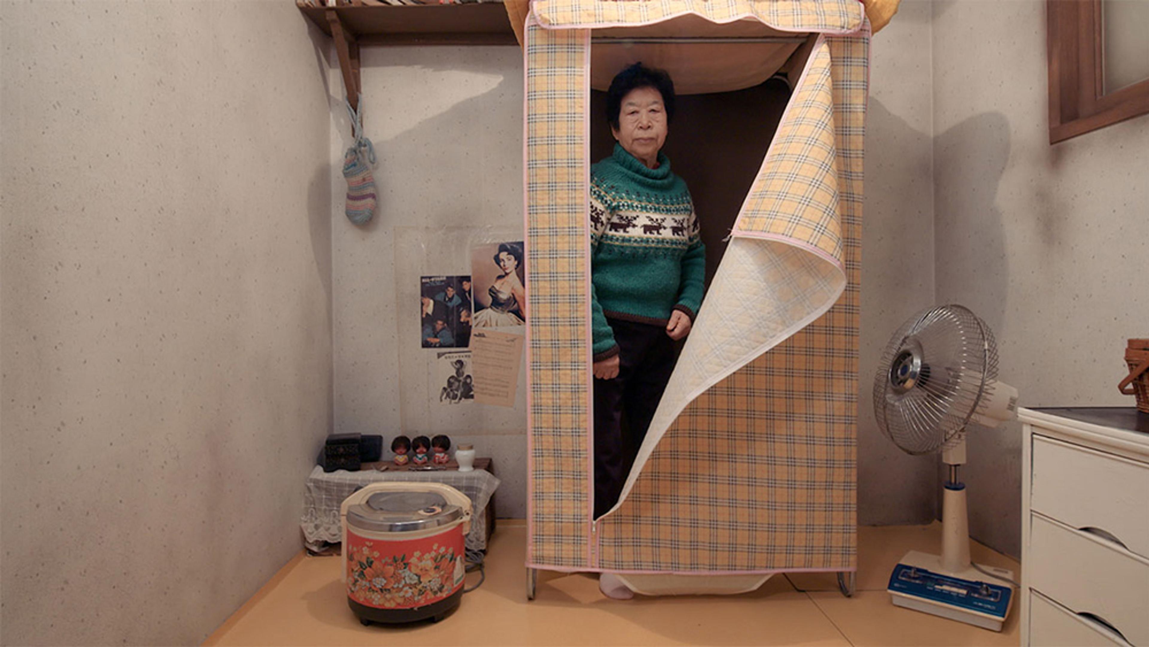 An elderly Asian woman standing inside a fabric wardrobe in a small room with vintage items, a fan, and posters on the wall.