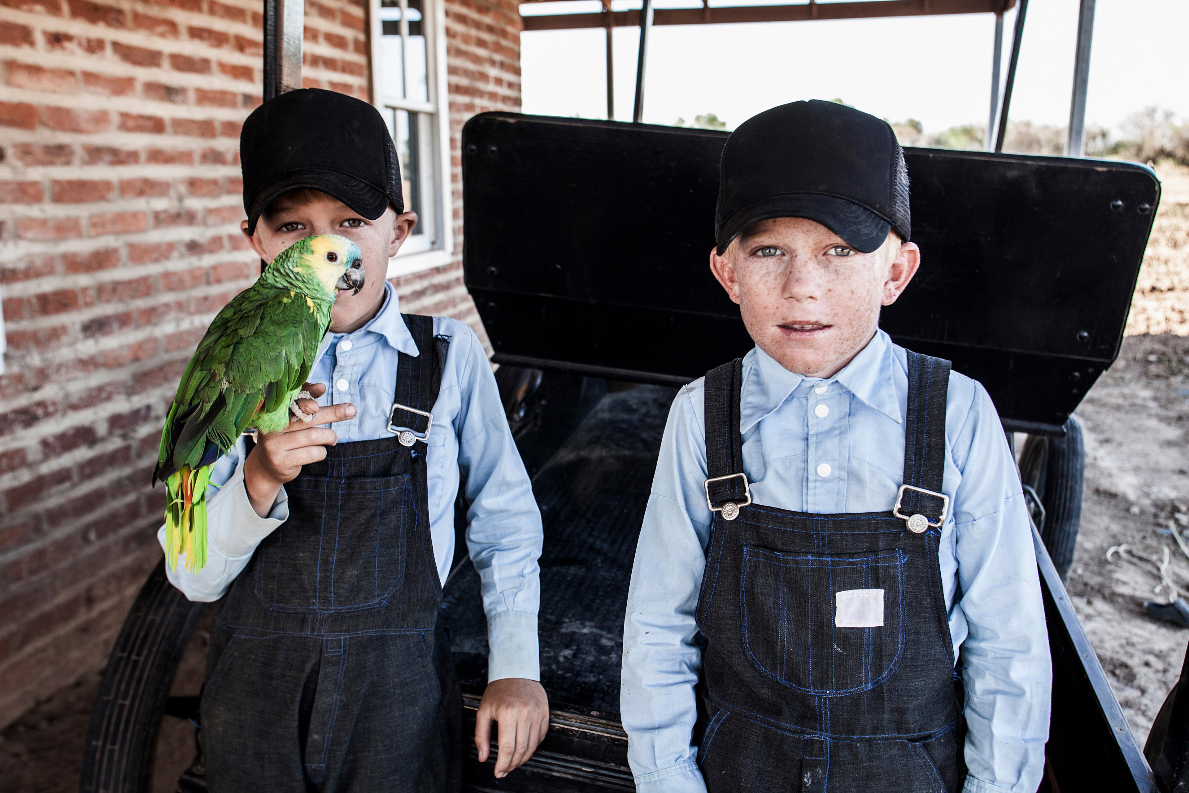 Photo of two boys in blue shirts and caps; one holds a green parrot. They stand beside a brick wall outside.