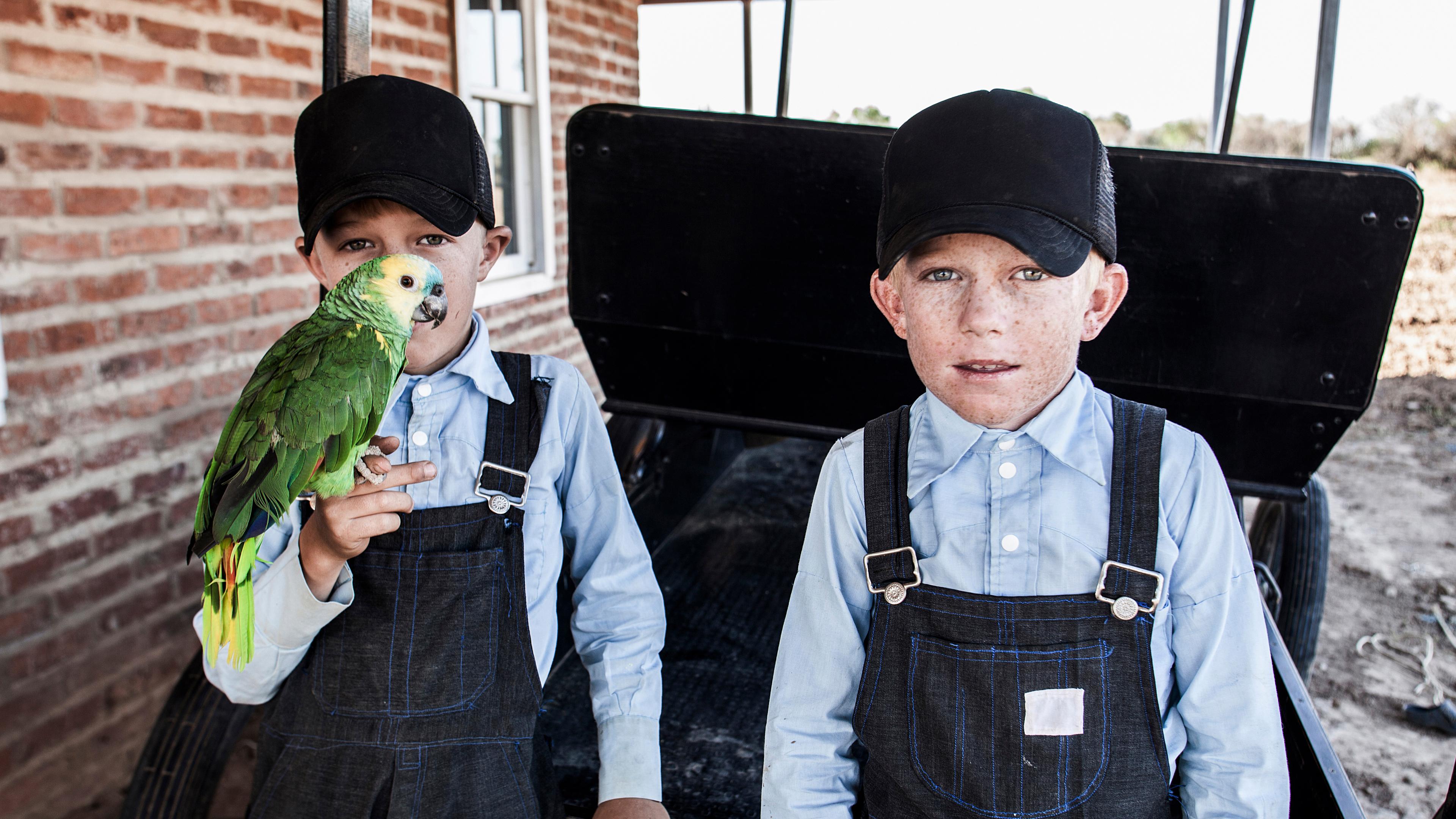 Photo of two boys in blue shirts and caps; one holds a green parrot. They stand beside a brick wall outside.