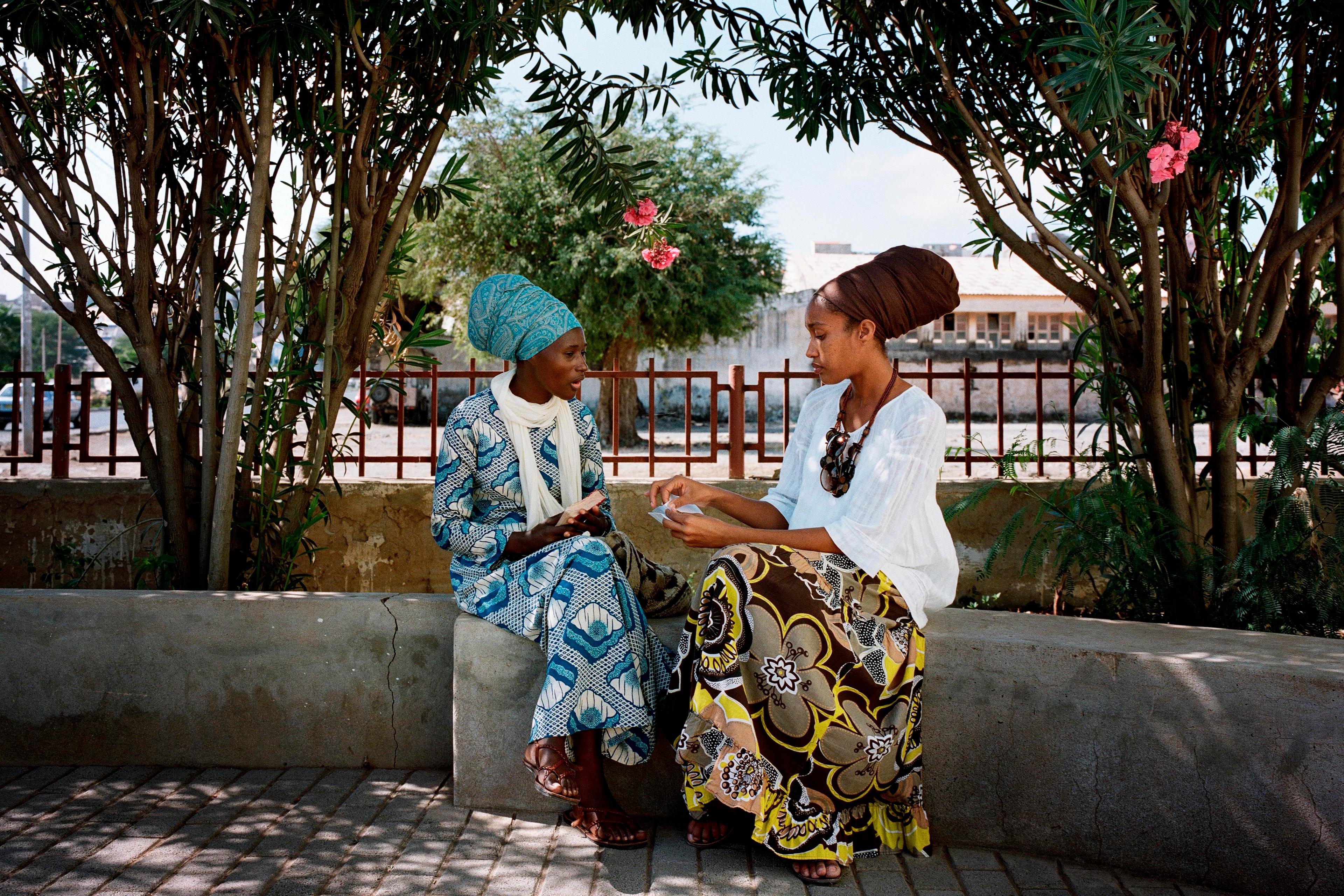 Photo of two women in colourful attire sitting on a bench under trees in a sunny outdoor setting, engaged in conversation.