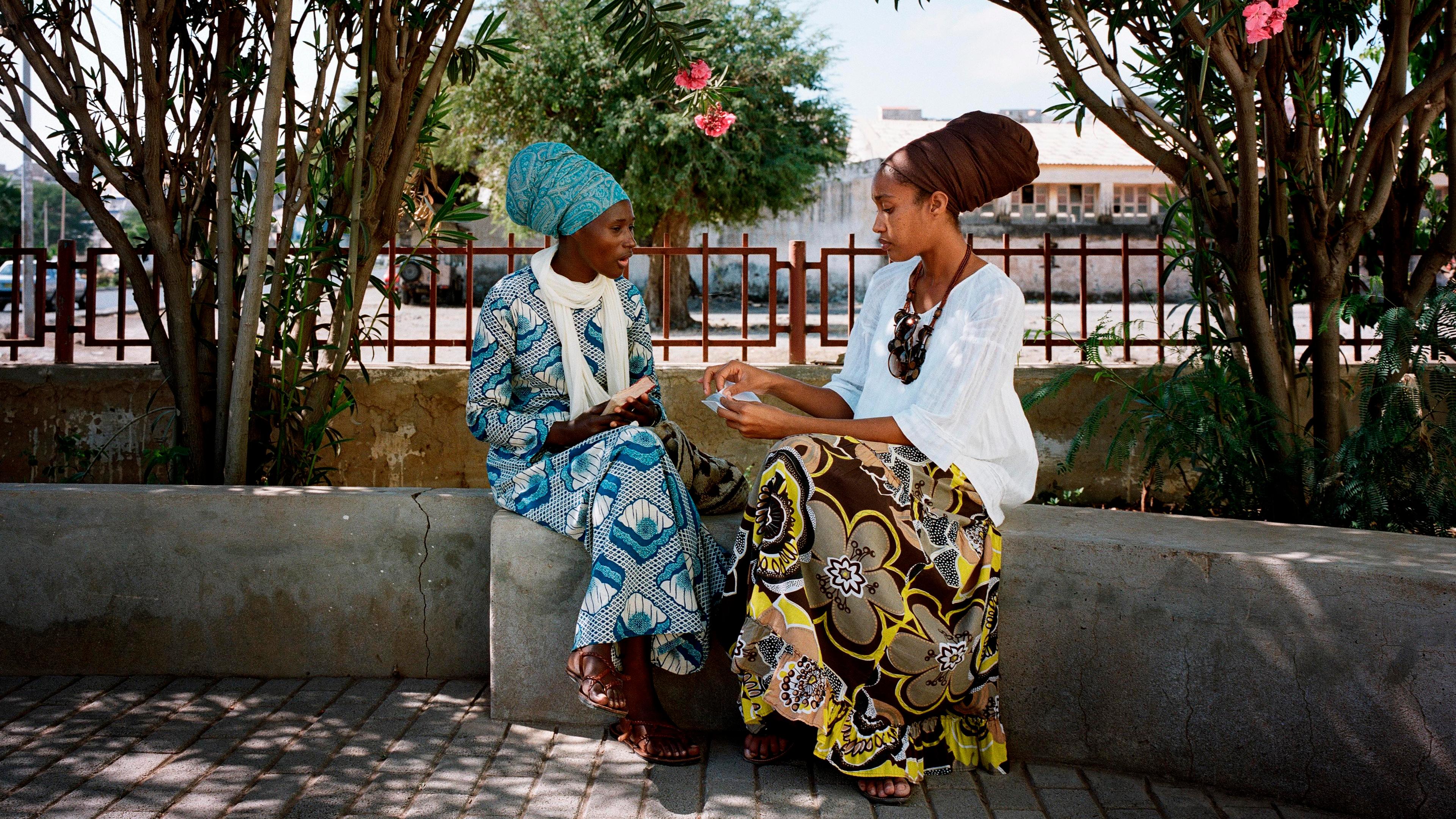 Photo of two women in colourful attire sitting on a bench under trees in a sunny outdoor setting, engaged in conversation.