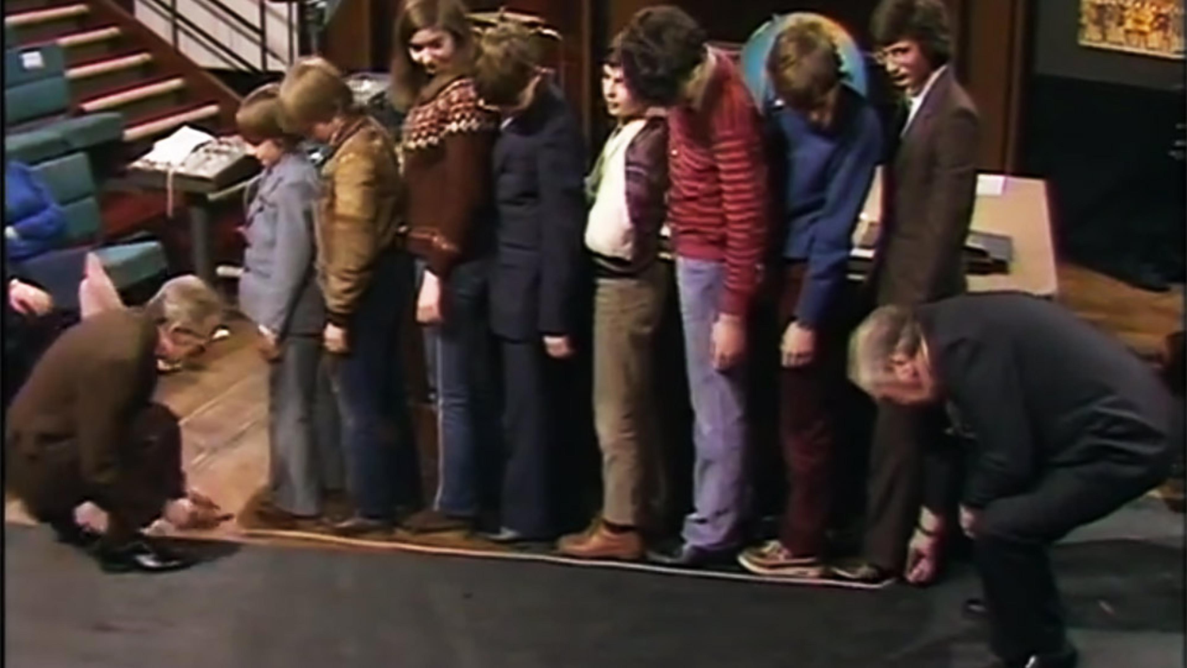 A group of people standing in a line on stage with two men measuring them.