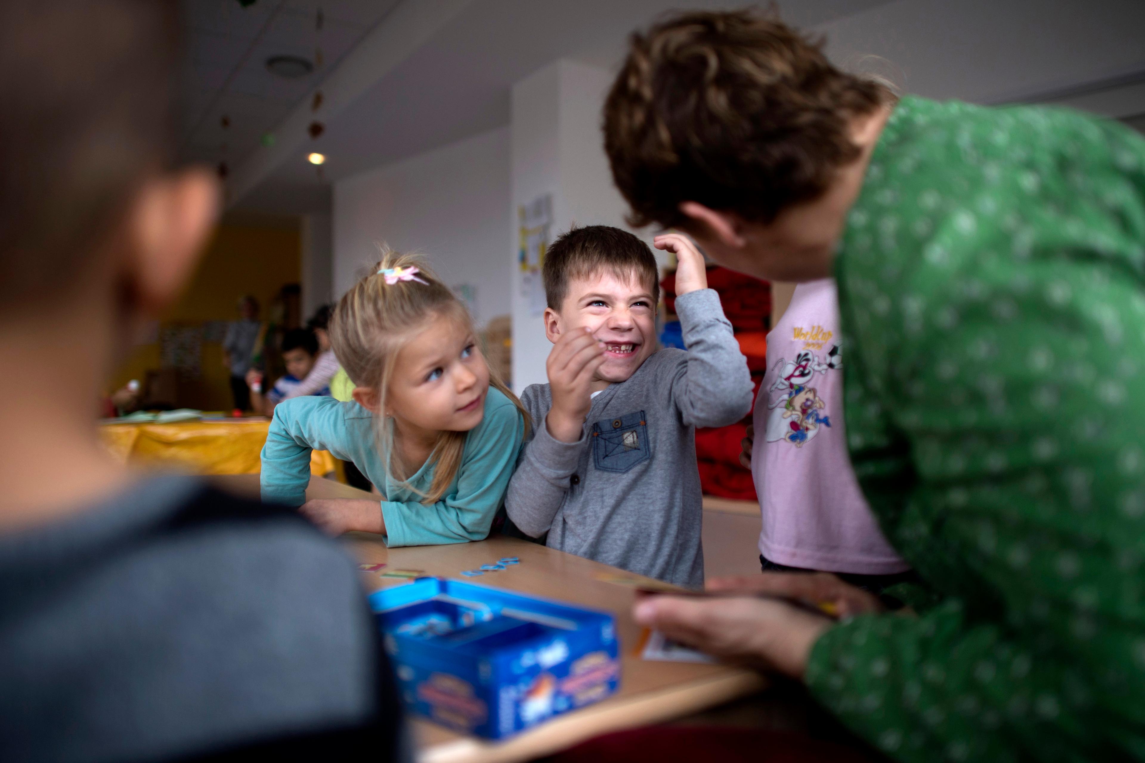 Photo of children in a classroom interacting with a teacher, sitting at a table with a board game.