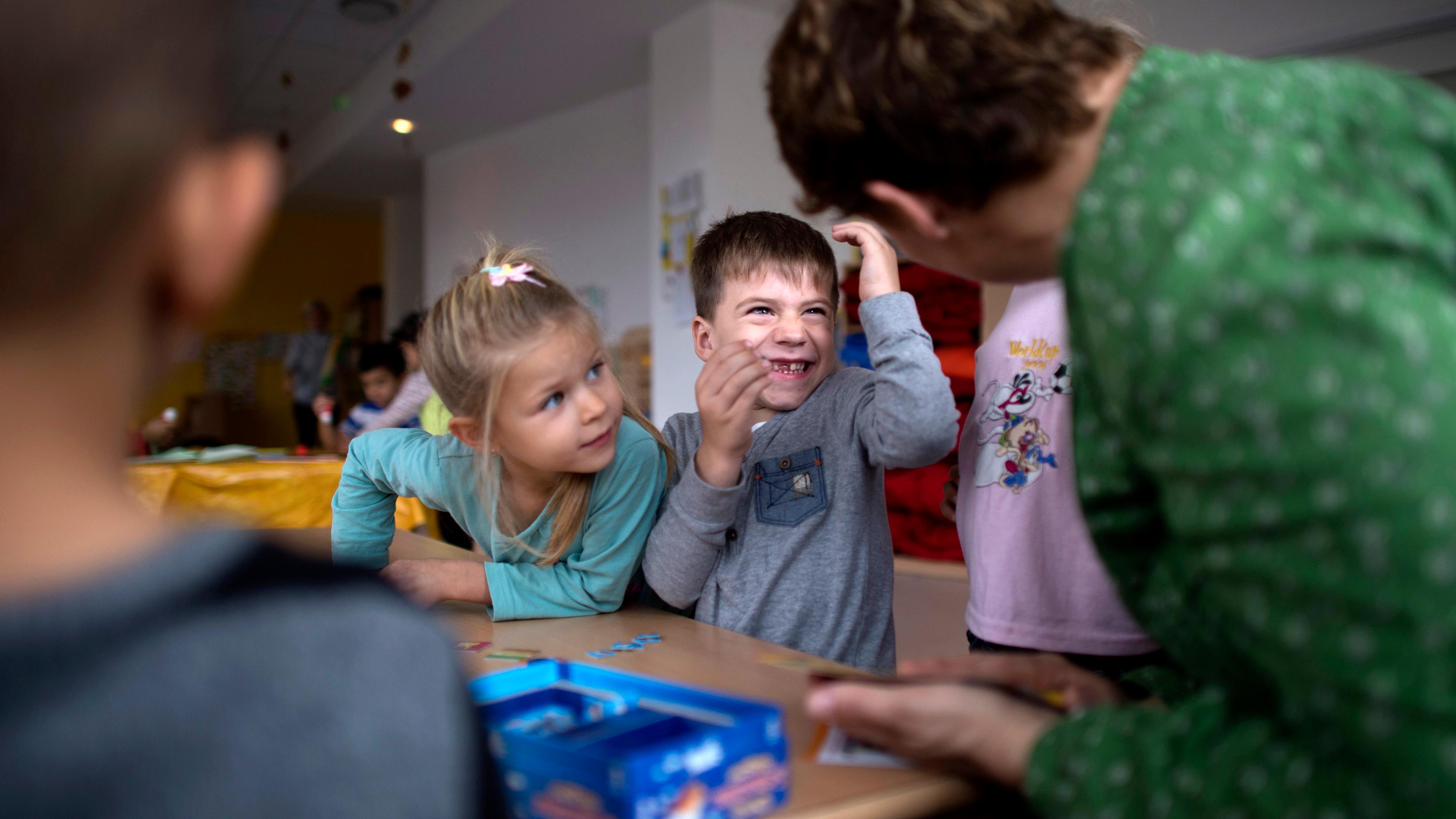 Photo of children in a classroom interacting with a teacher, sitting at a table with a board game.