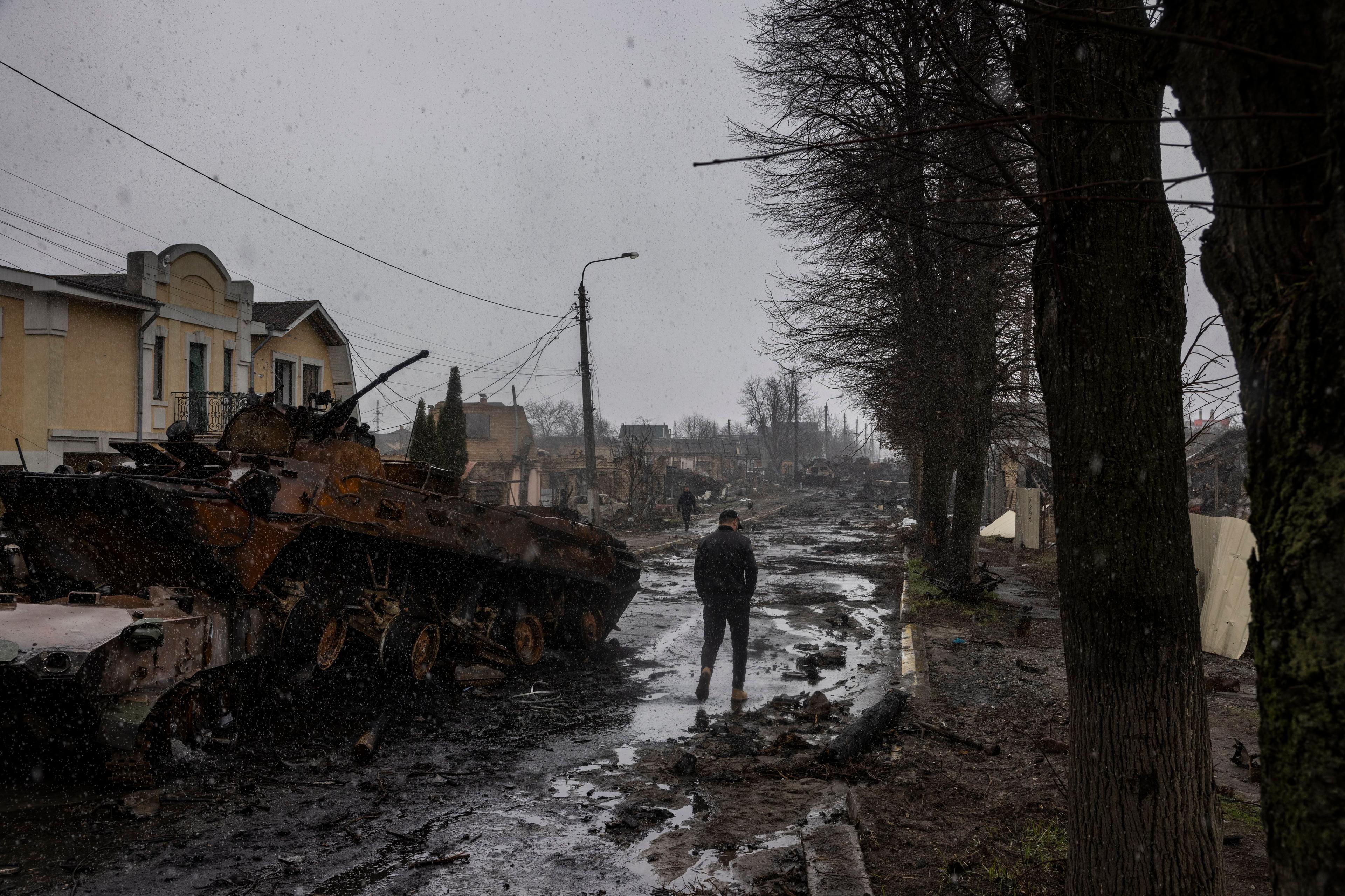 Photo of a war-torn street with a destroyed tank, damaged buildings and a person walking on a muddy path during snowfall.