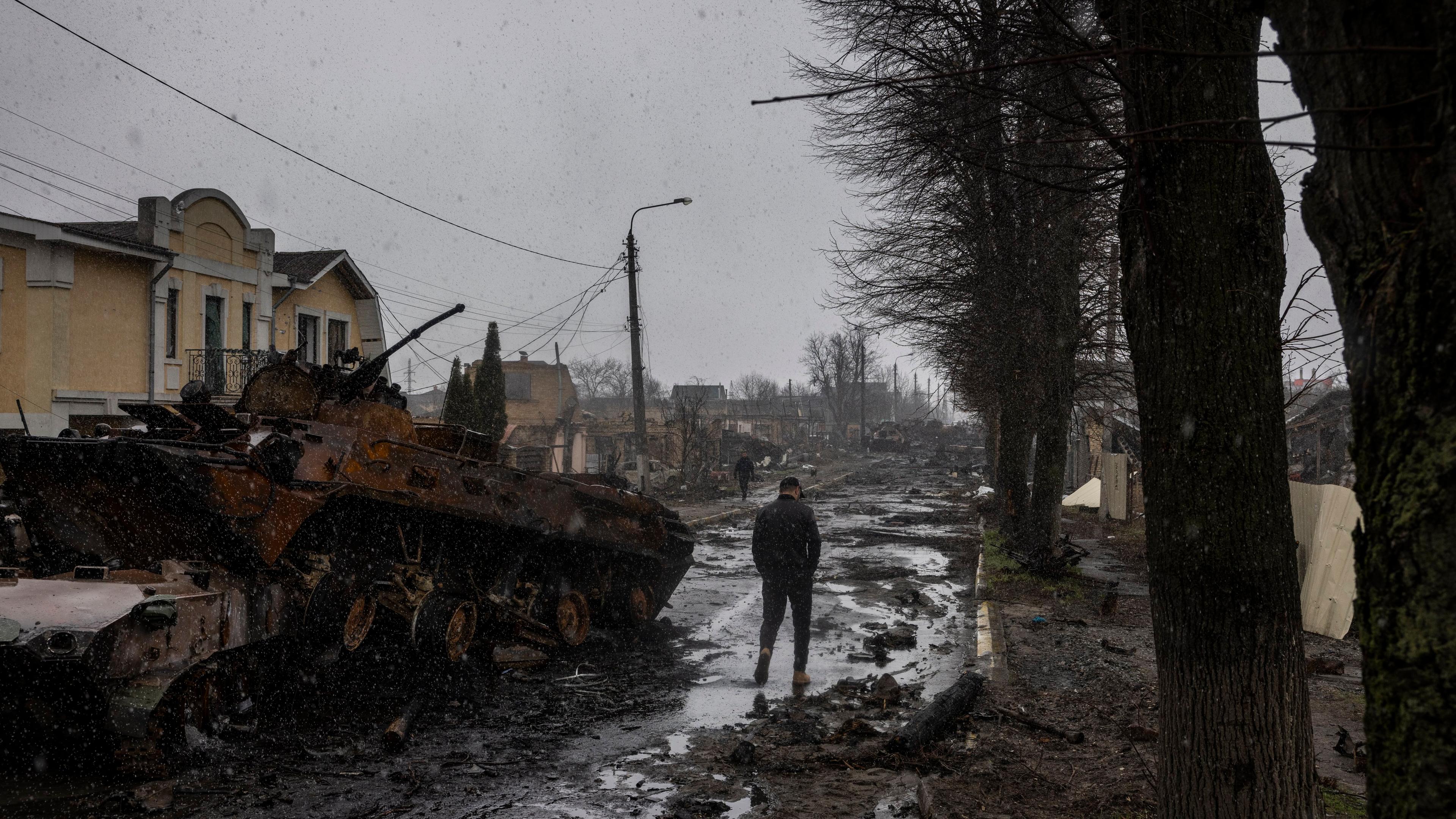 Photo of a war-torn street with a destroyed tank, damaged buildings and a person walking on a muddy path during snowfall.