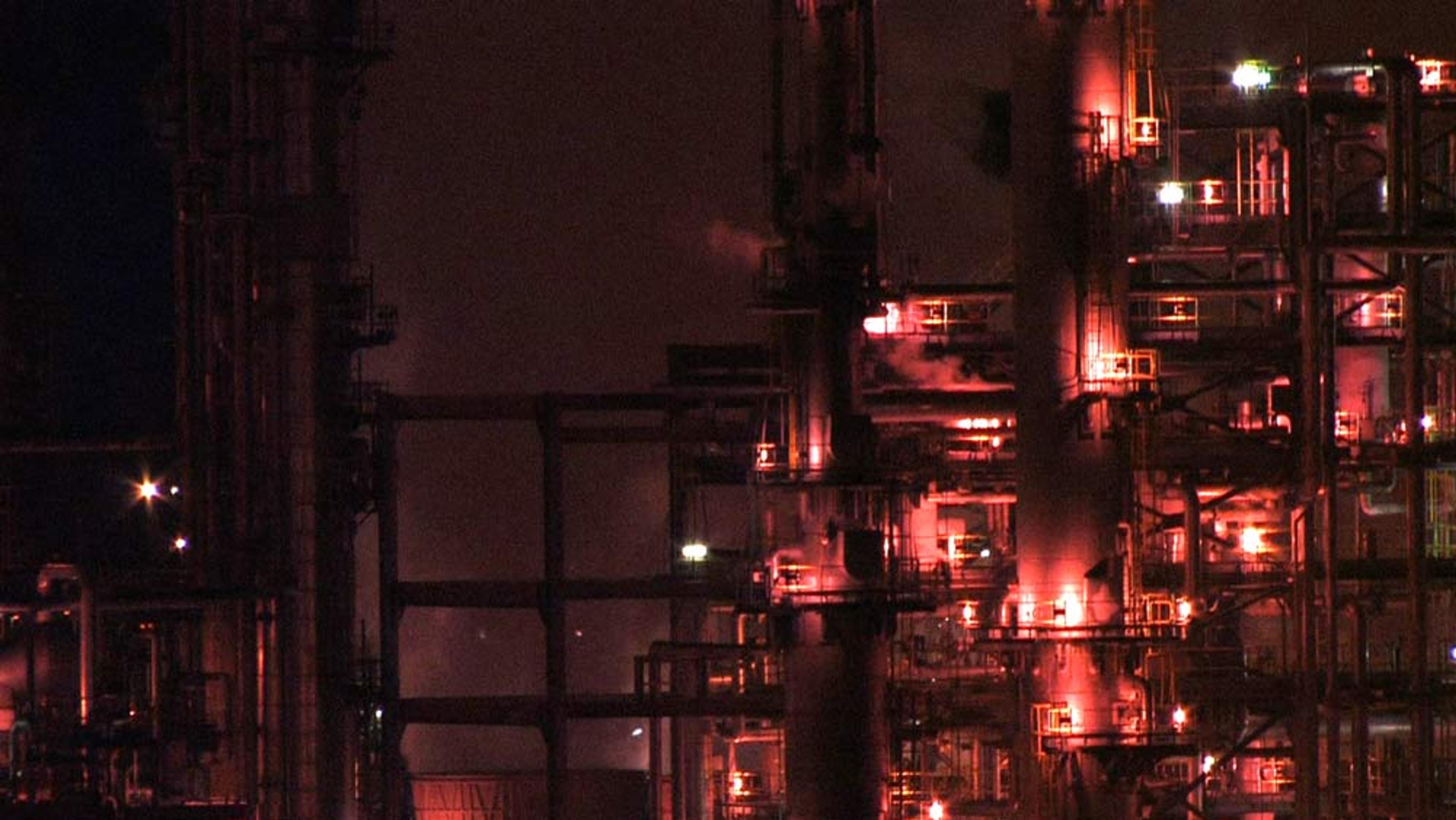 An industrial facility at night with lit structures and piping, emitting smoke and illuminated by red and orange lights.