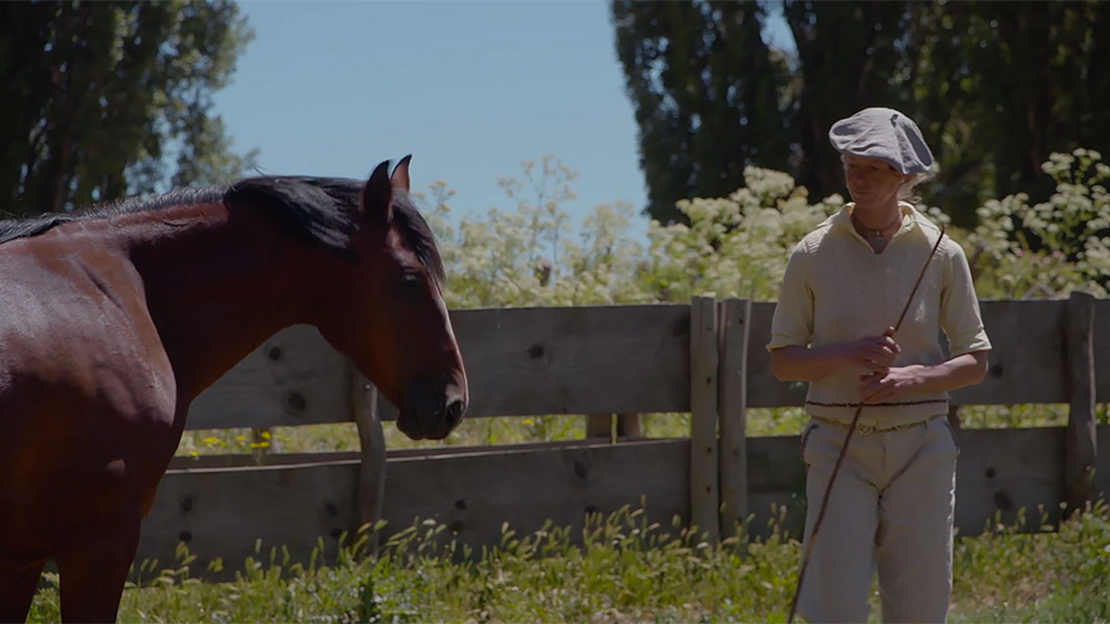 A woman holding a stick standing next to a brown horse in an outdoor setting with wooden fencing and greenery.