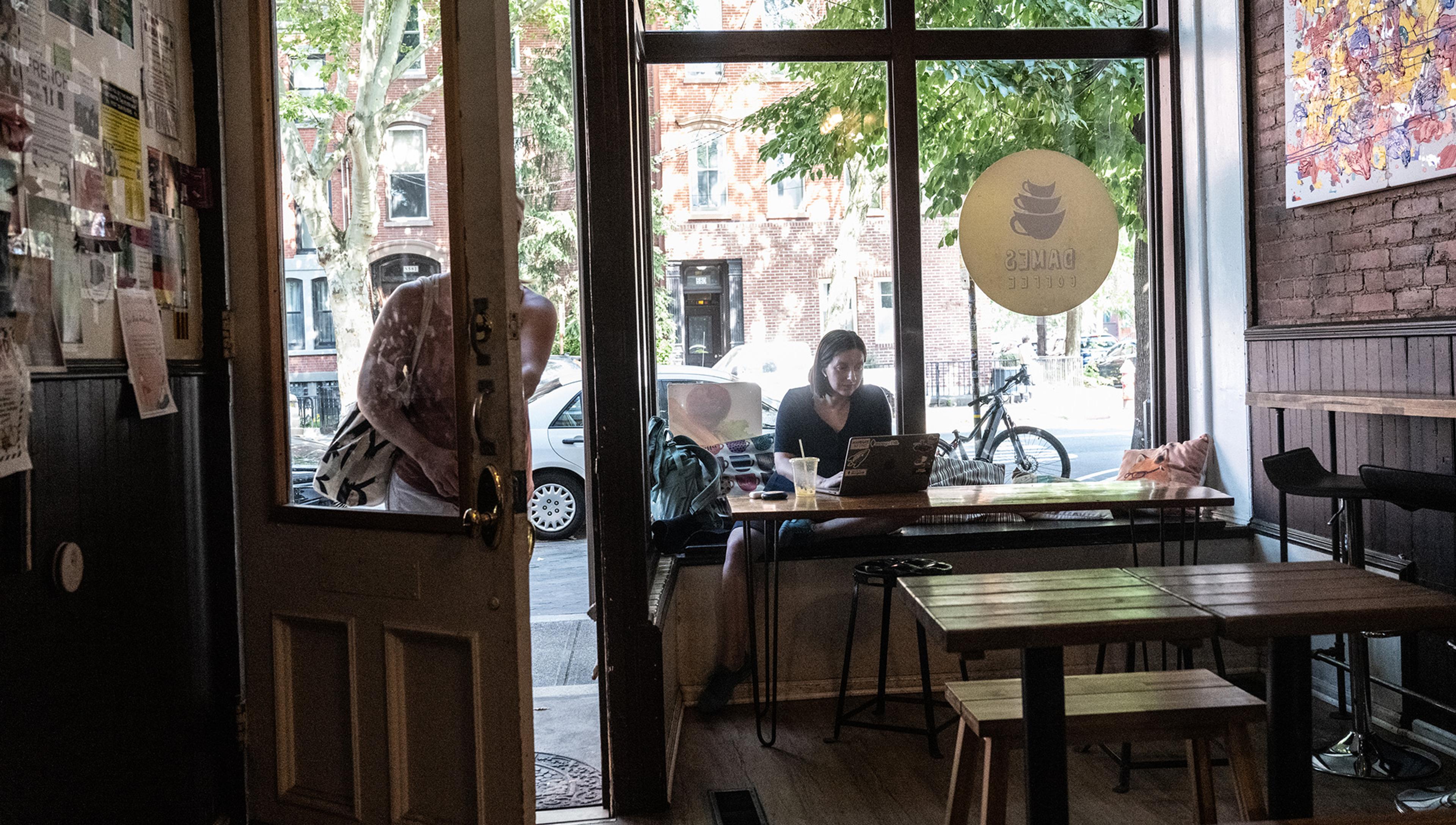 Photo of a café interior showing a person working on a laptop inside by the window while another person is entering through the door.