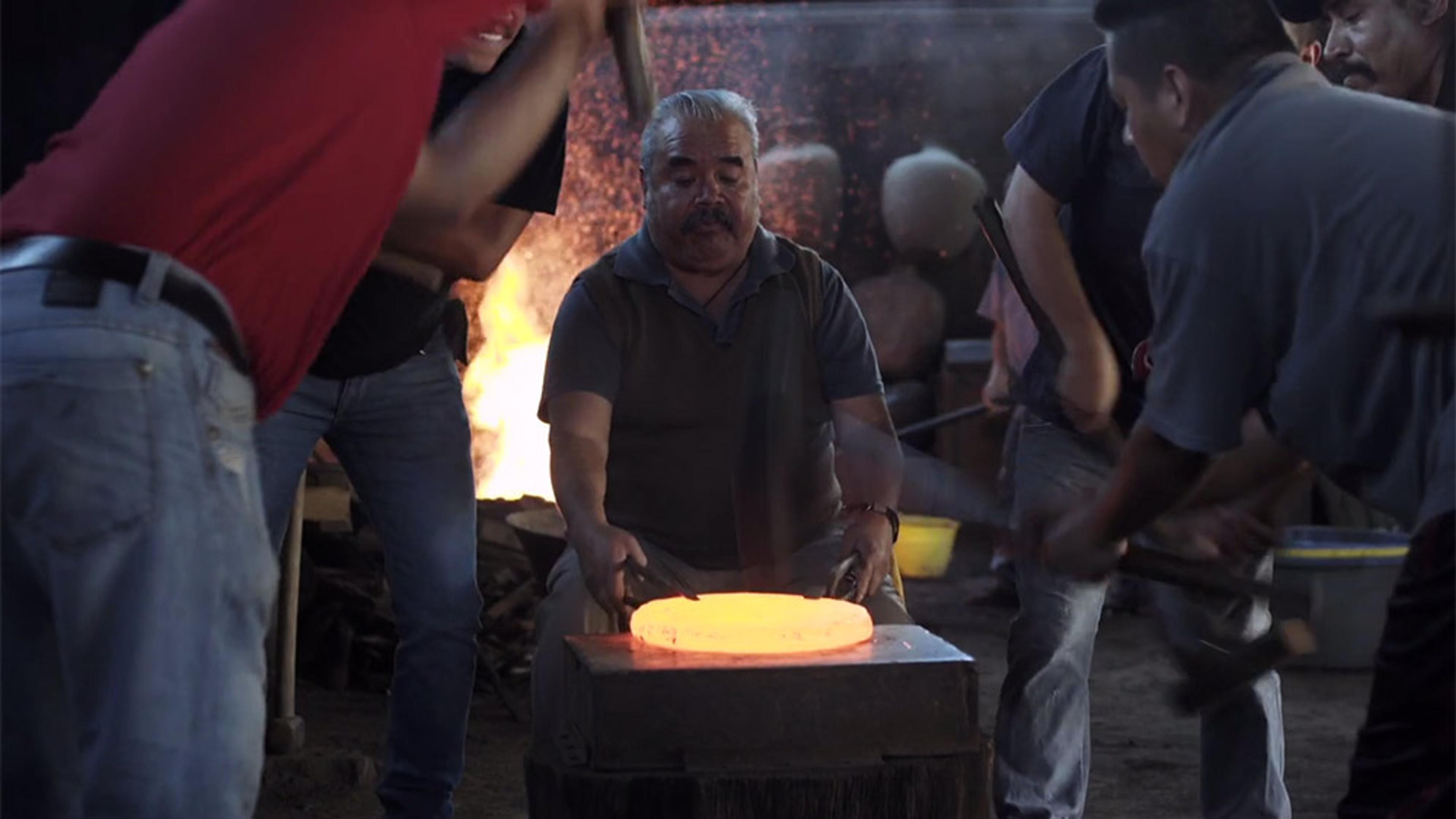 Blacksmiths hammering a glowing piece of metal while another man holds it steady near a forge with flames in the background.