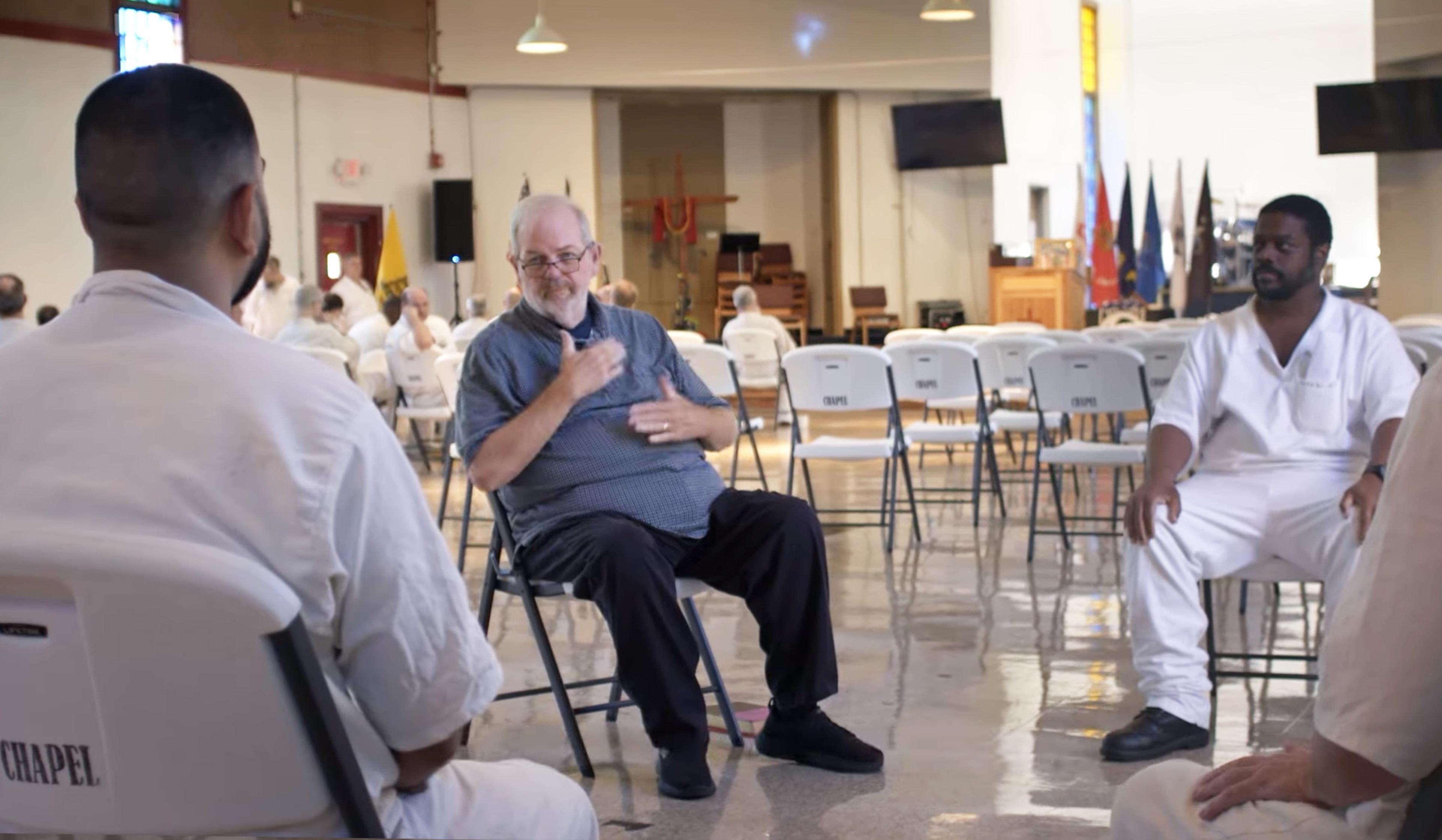 Several deaf inmates sit on chairs in a circle, engaged in an ASL conversation. Room has white walls, white chairs, flags and a podium in the background.