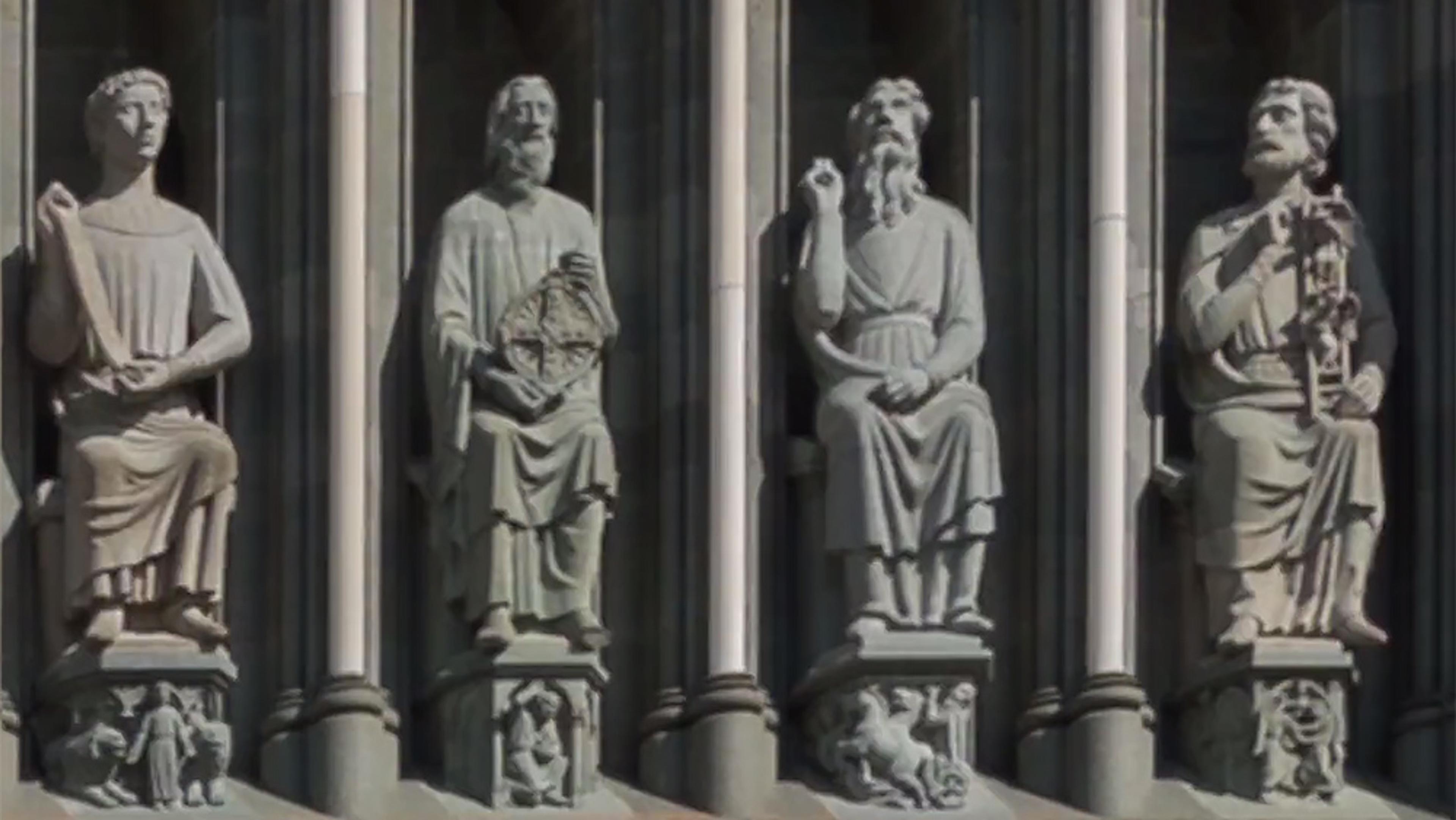 Four stone statues of seated figures on a building exterior, each holding different objects and flanked by decorative columns.