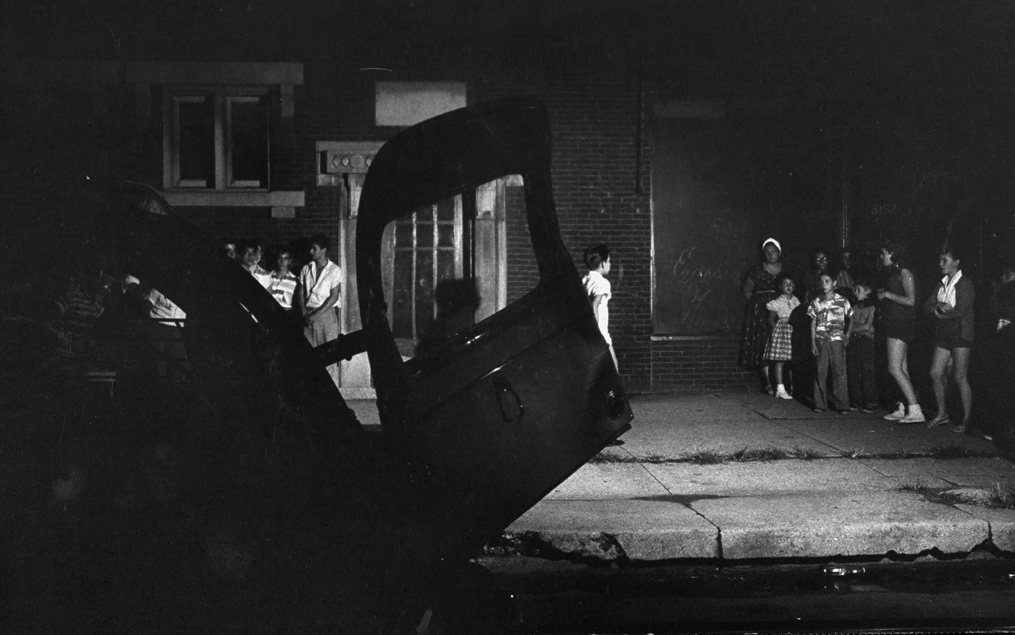 Vintage photo of a night scene with a large vehicle in the foreground and a group of people gathered on a pavement in front of a building.