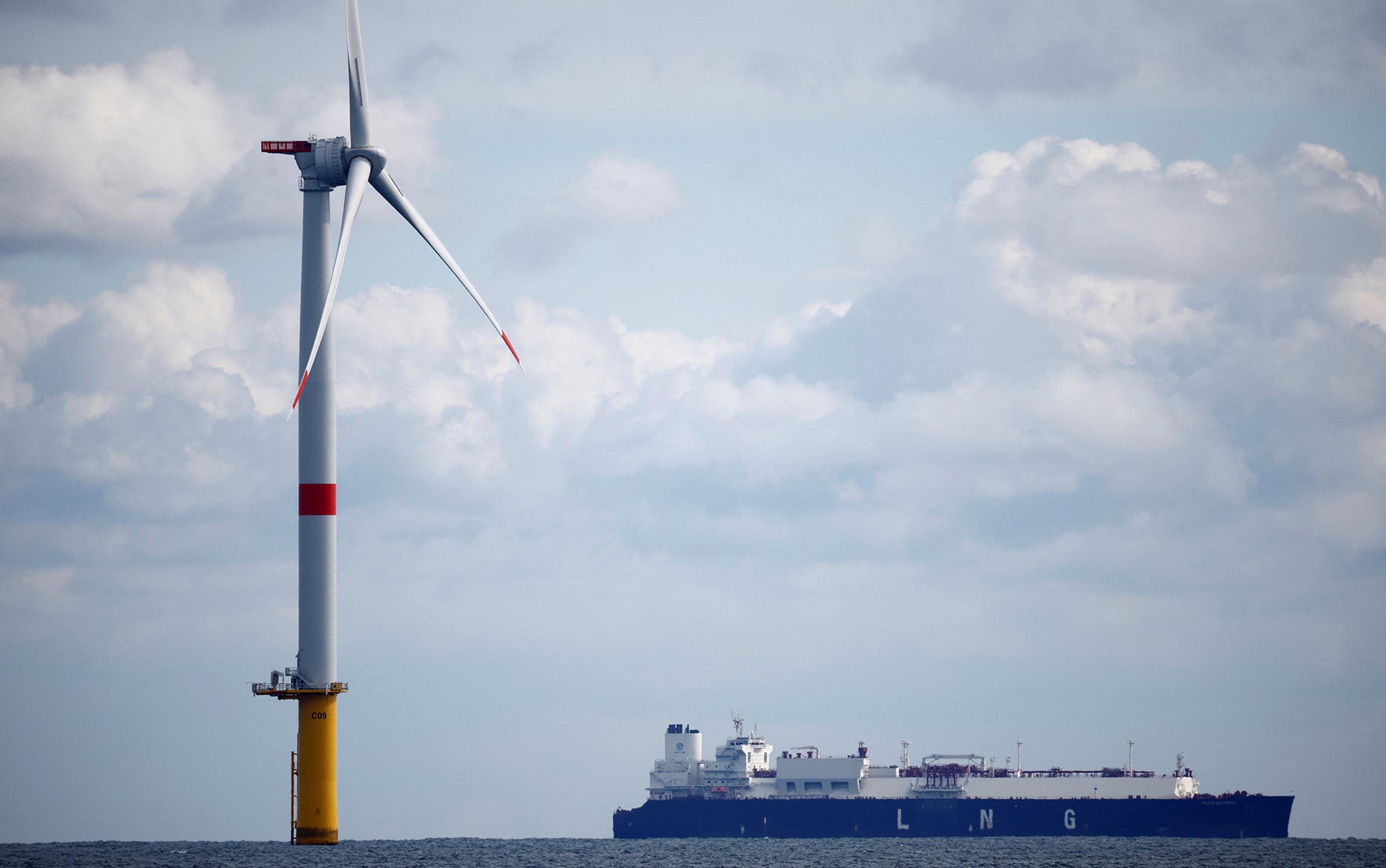 Image of an offshore wind turbine in the sea with a large LNG ship in the background under a partly cloudy sky in calm waters.