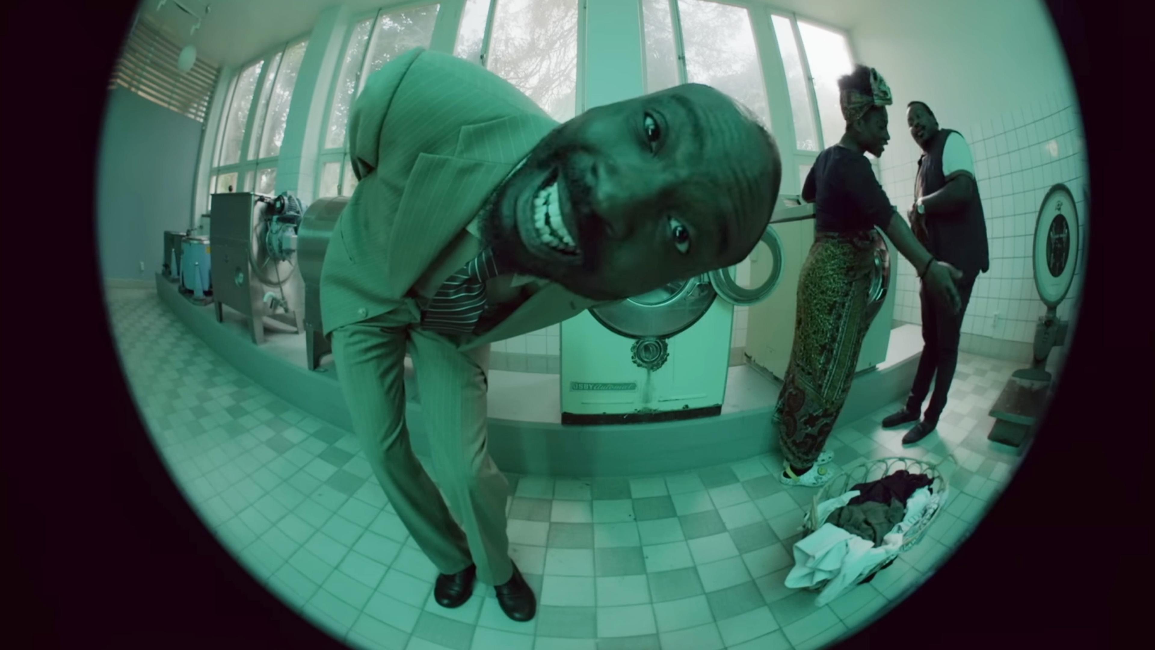 Fisheye photo of a man in a suit leaning close to camera in a laundrette, with two people talking in the background.
