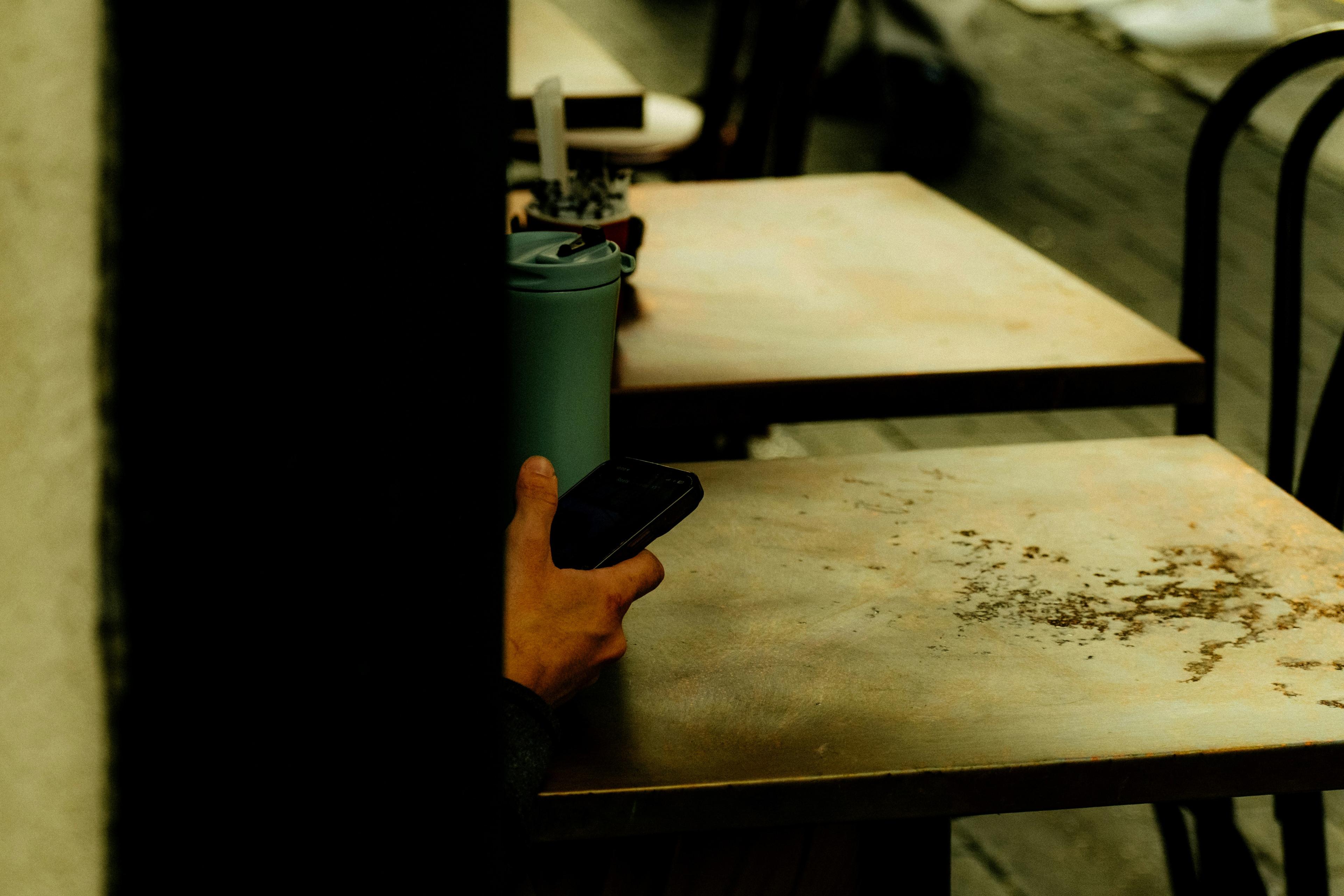 A person partially hidden holding a smartphone and a green cup at a table in a café setting.