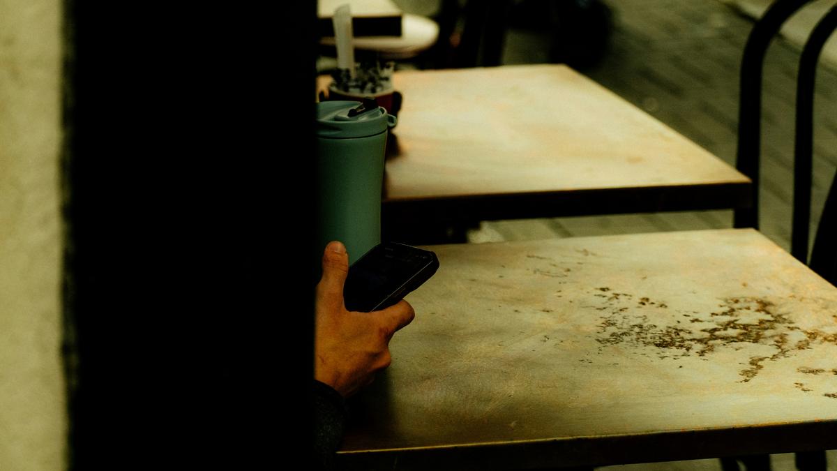 A person partially hidden holding a smartphone and a green cup at a table in a café setting.