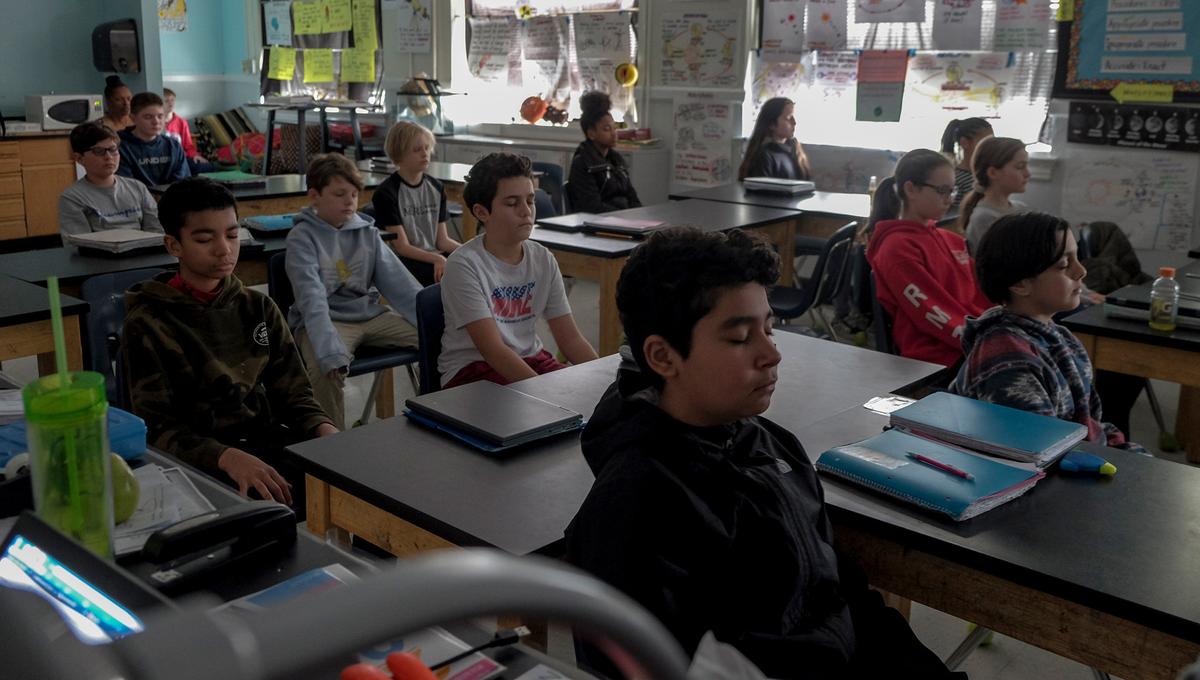 Classroom of middle-school students sitting at desks with eyes closed, appearing to be meditating or focusing quietly.