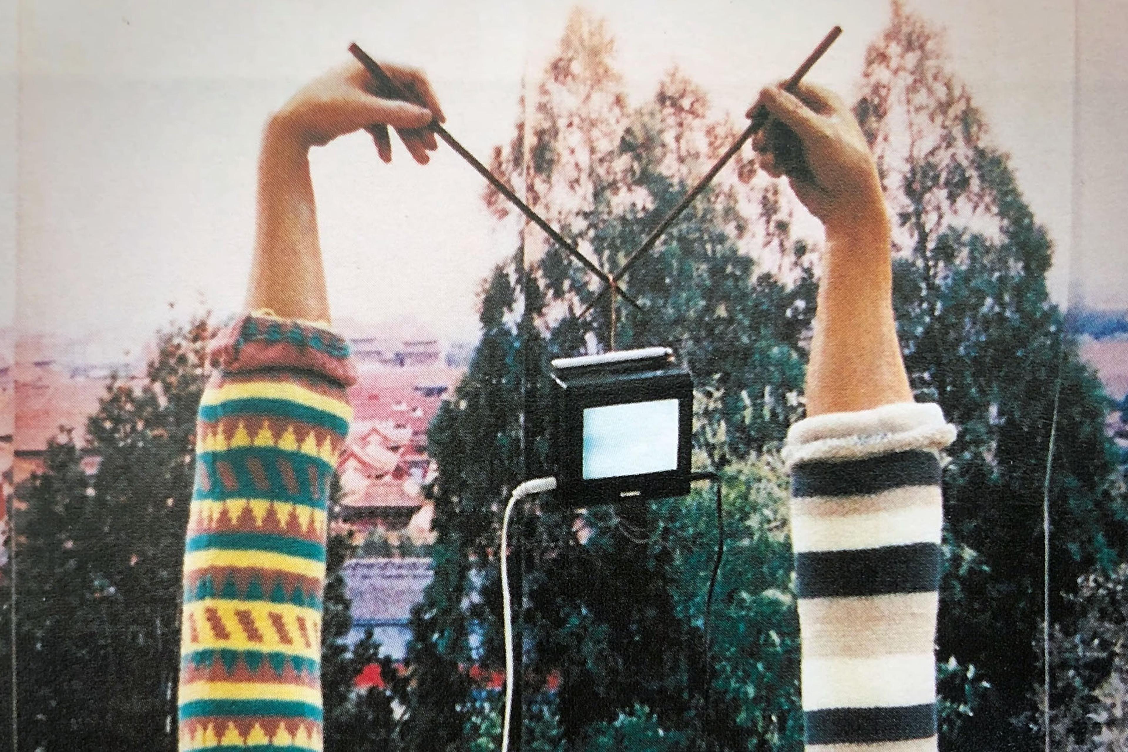 Two arms in colourful jumpers hold chopsticks that hold a small TV. Trees and rooftop scenery are visible in the background.