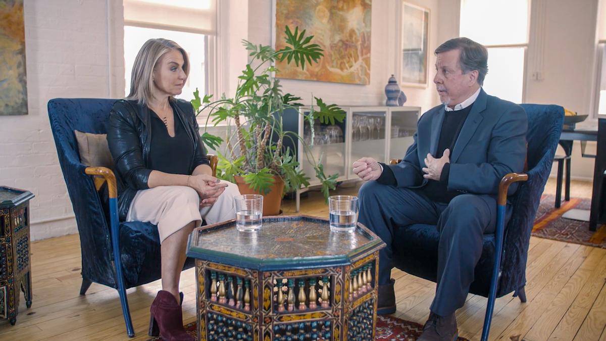 A woman and a man converse while seated on chairs in a room with plants. A decorative table with glasses is between them.