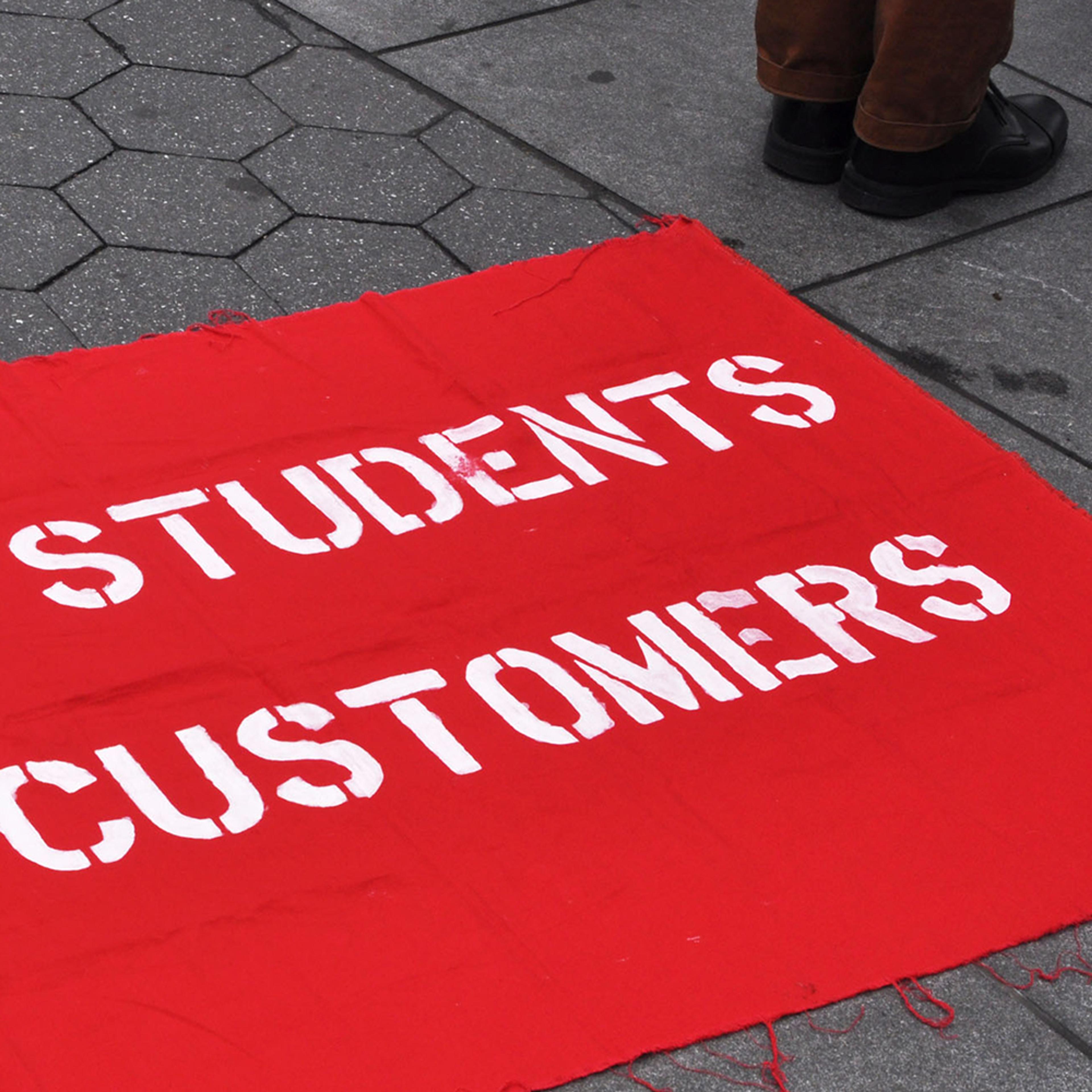 A red cloth on the ground with the words “STUDENTS CUSTOMERS” written in white. A person’s feet are visible nearby.