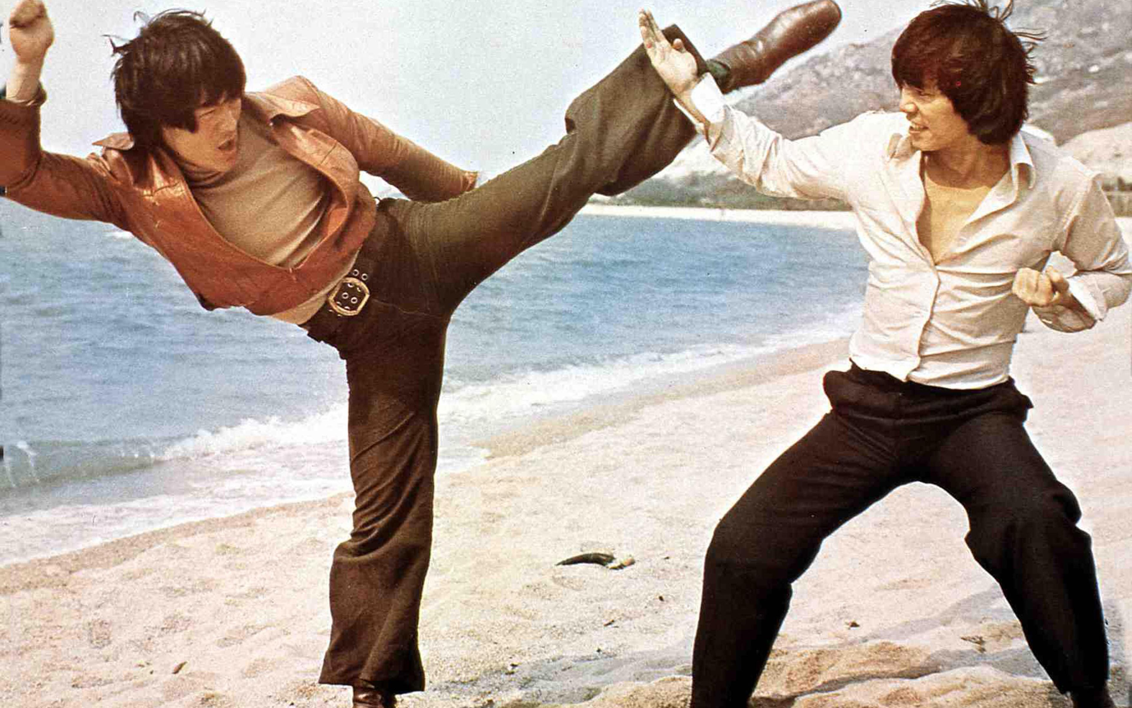 Two Asian film actors are fighting kung-fu style on the beach dressed in 1970s style casual clothes