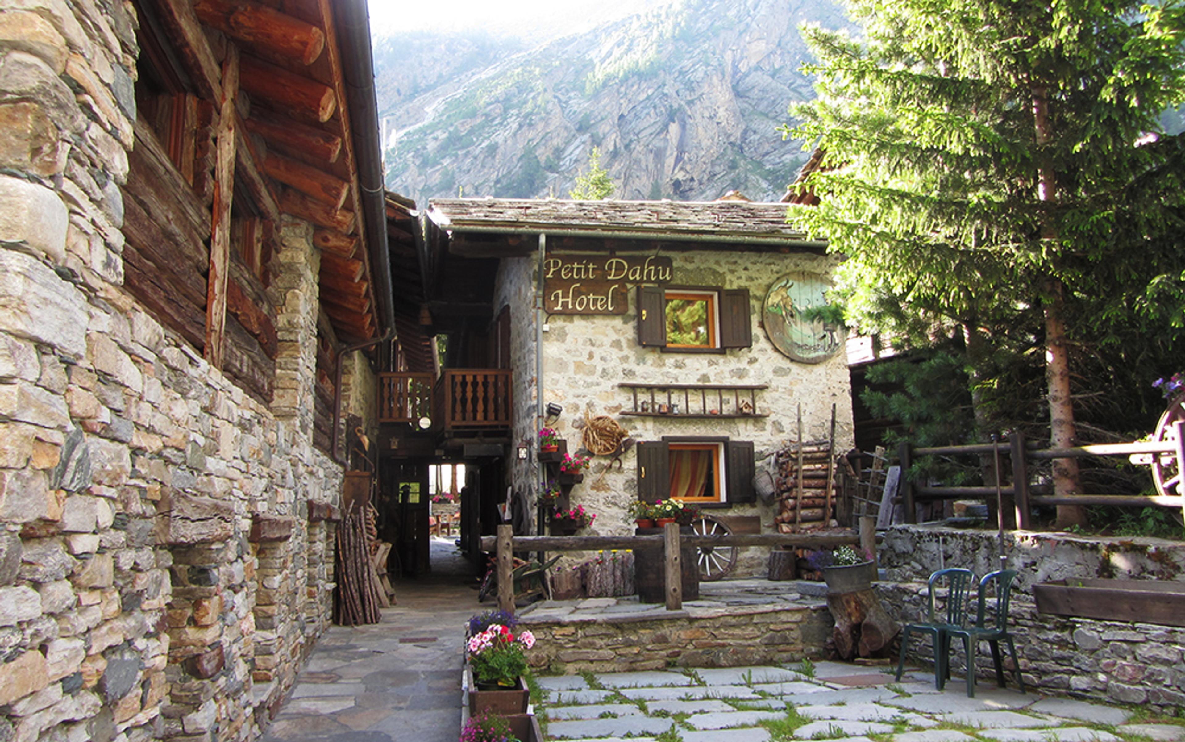 Arustic stone building with the sign “Petit Dahu Hotel”, surrounded by greenery and mountains.