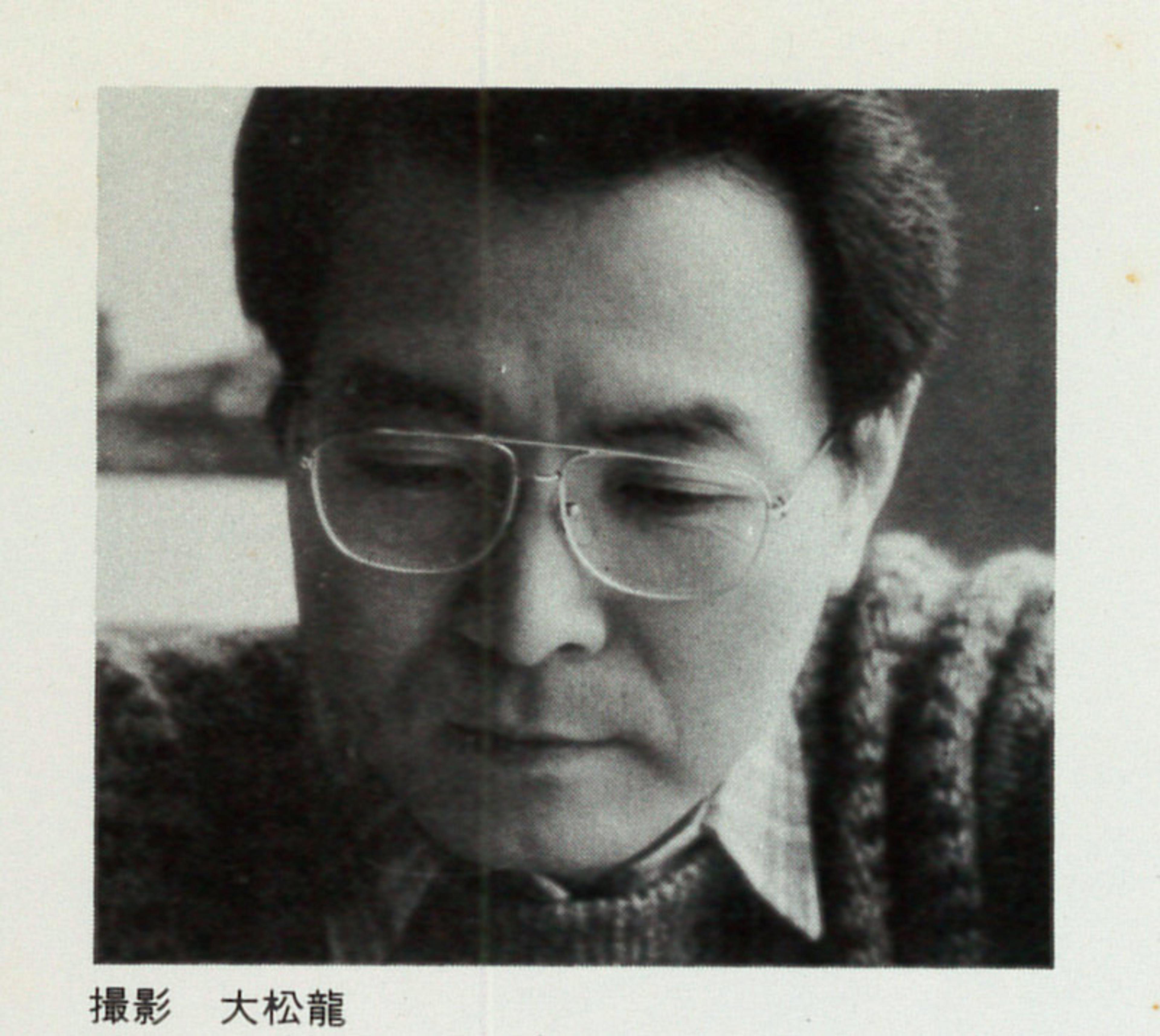 Black and white photo of a middle-aged man in glasses and a sweater looking down. There is Japanese text beneath the photo.