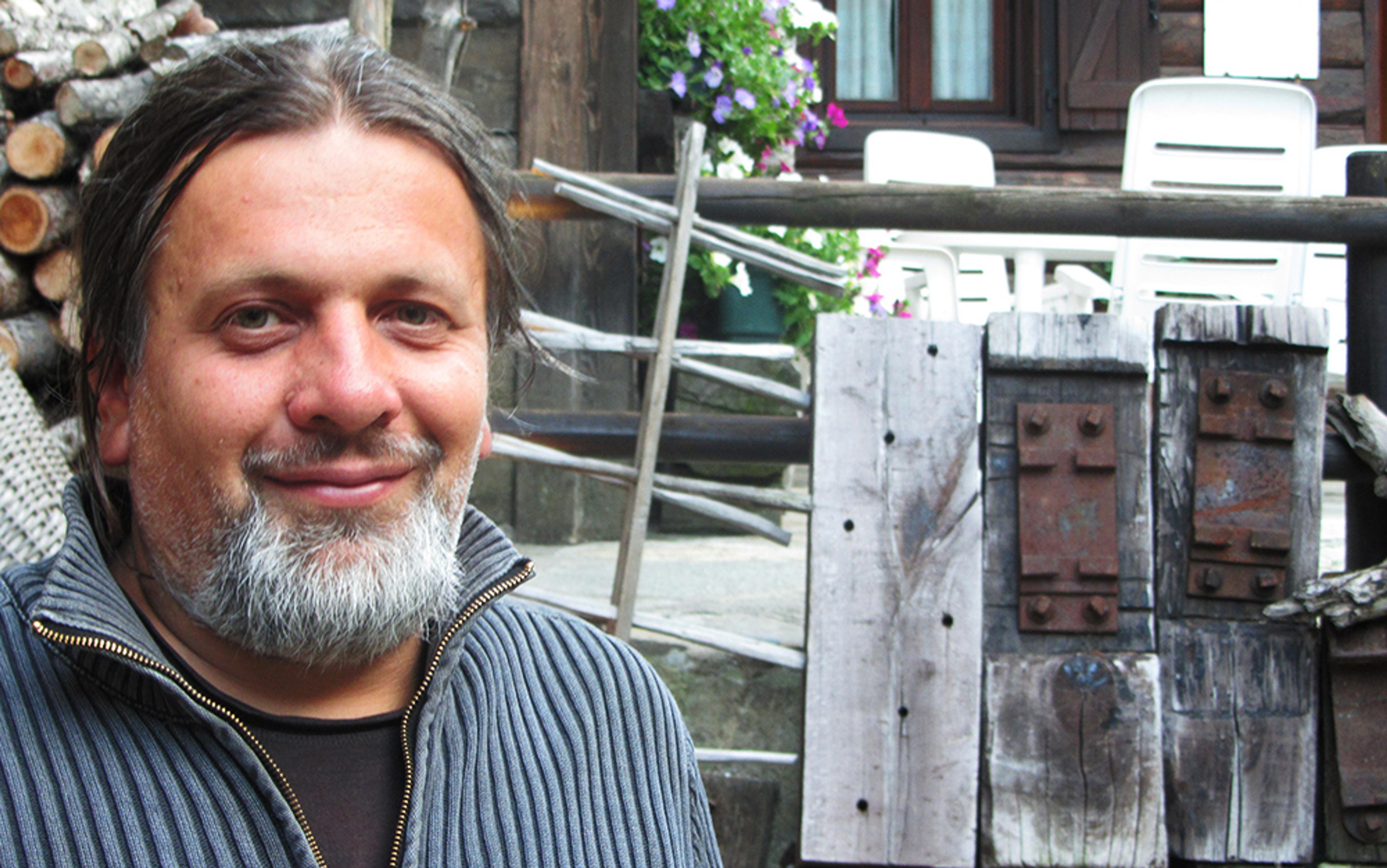 A man with long hair and a beard smiling outdoors with logs and wooden beams behind him in a rustic setting, photo.