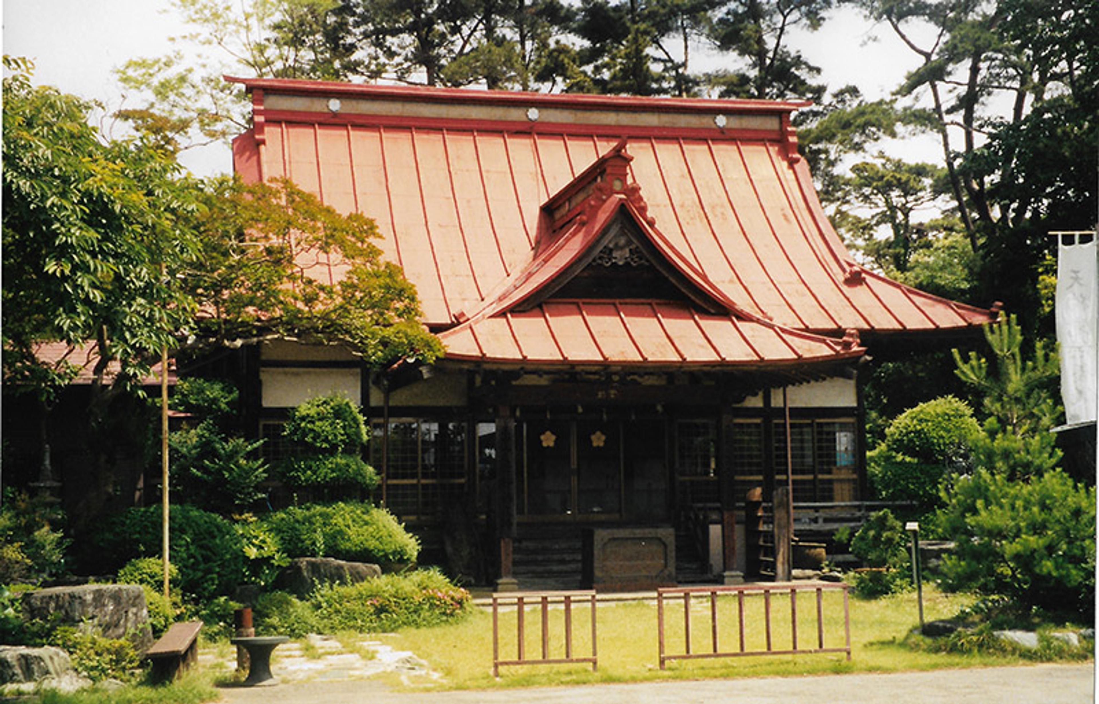 Photo of a Japanese temple with a steep red roof and dark wood features surrounded by greenery.