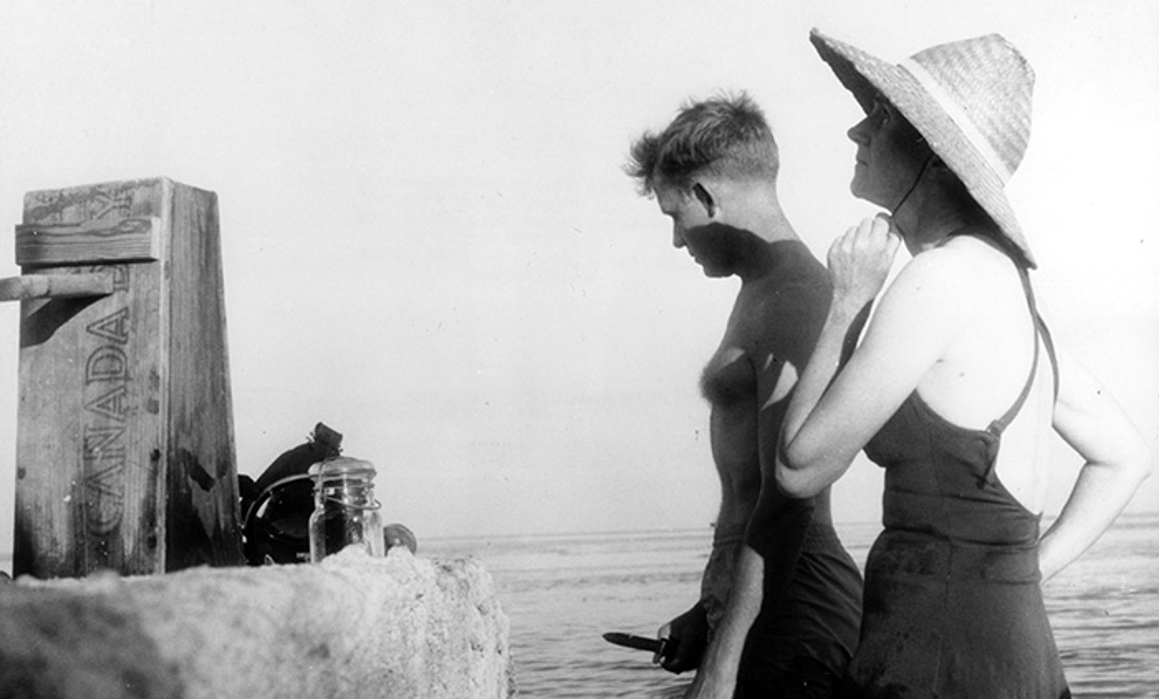 Vintage photo of a man and woman at the seaside. The woman wears a sunhat and the man holds an object. A box reads “CANADA”.
