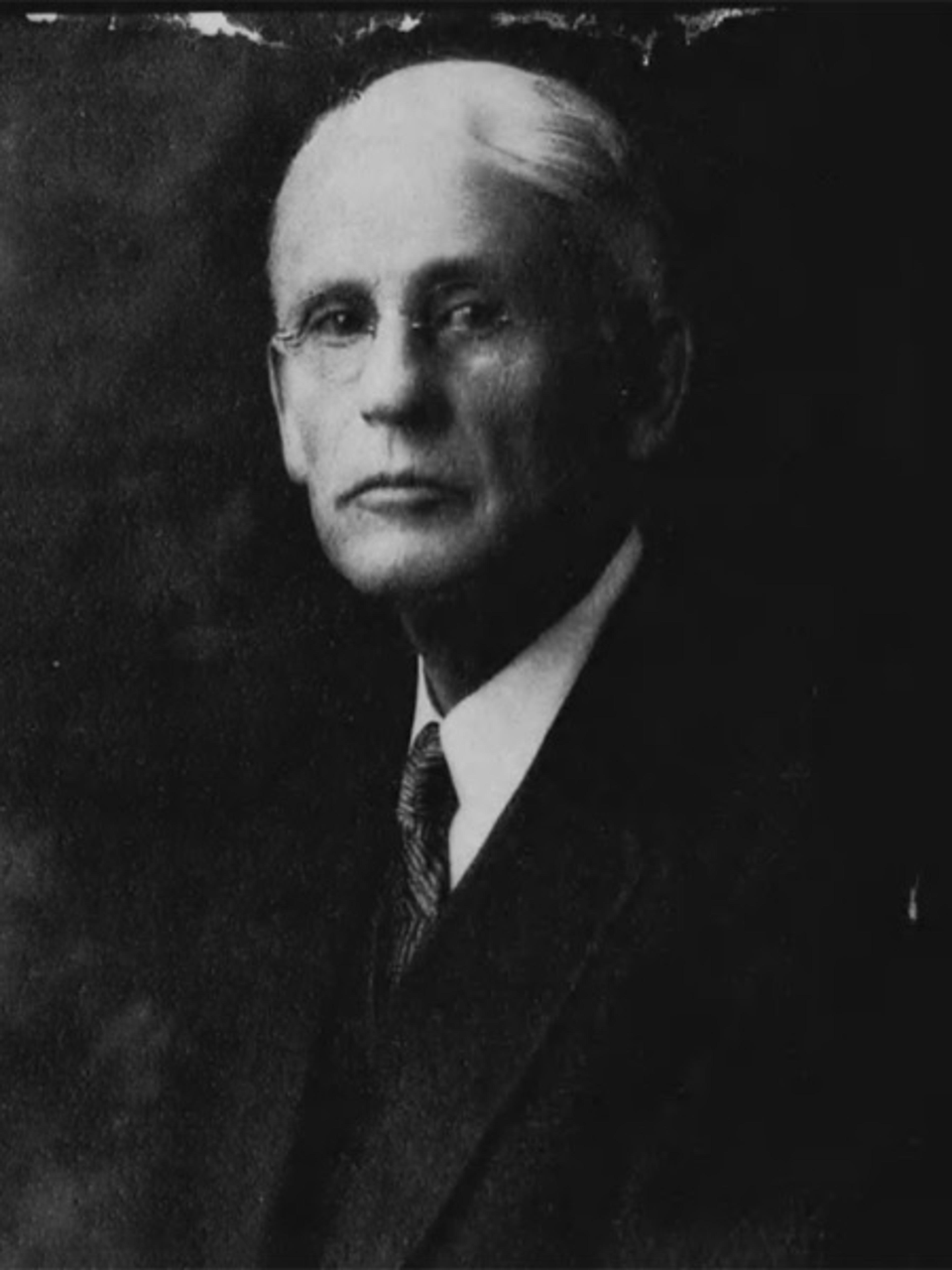 Black-and-white photo of an elderly man with glasses in a suit and tie, looking towards the camera with a serious expression.
