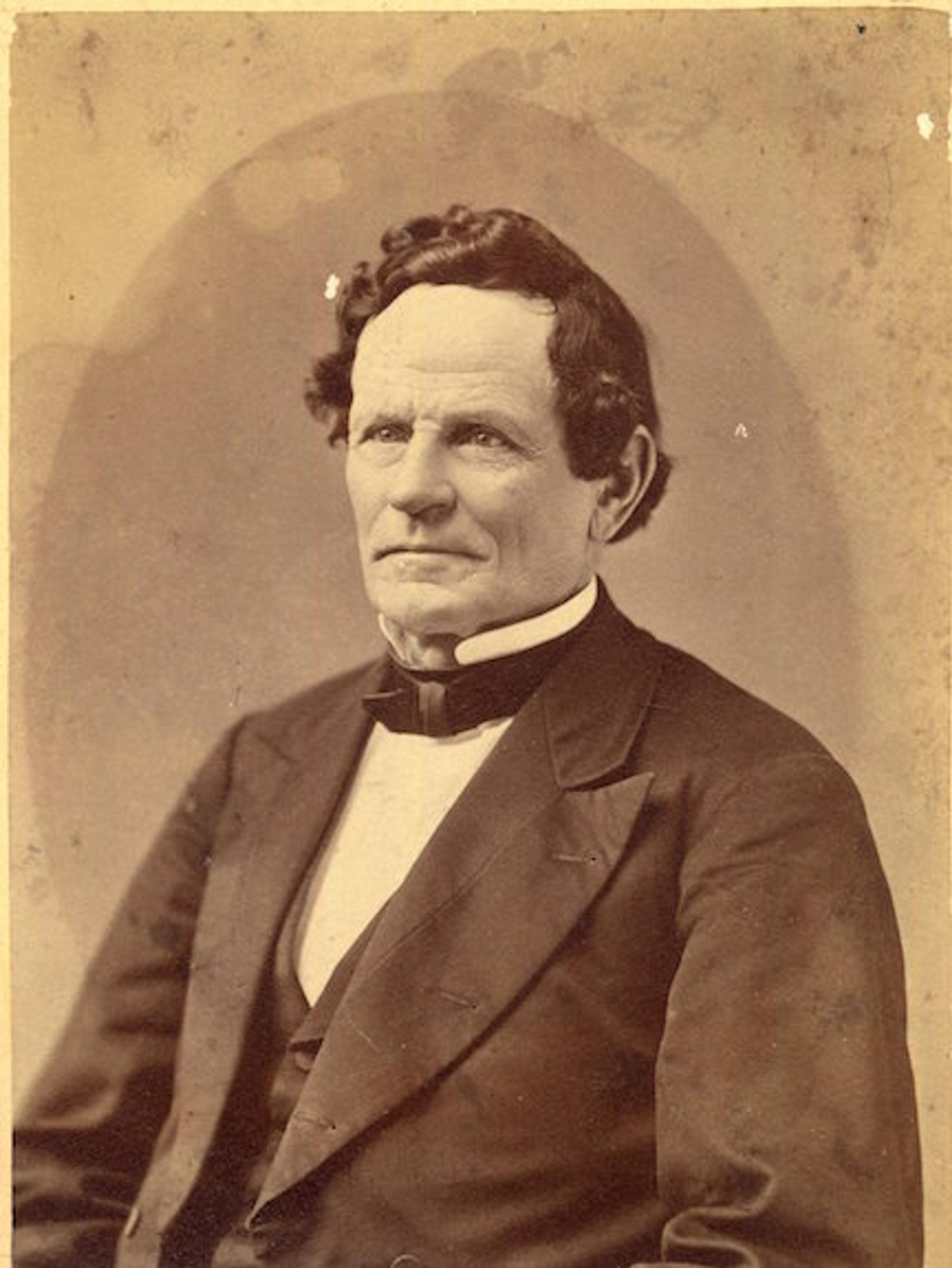 Sepia-toned photograph of an older man in a suit with a bowtie, sitting and looking slightly to the side, circa 19th century.