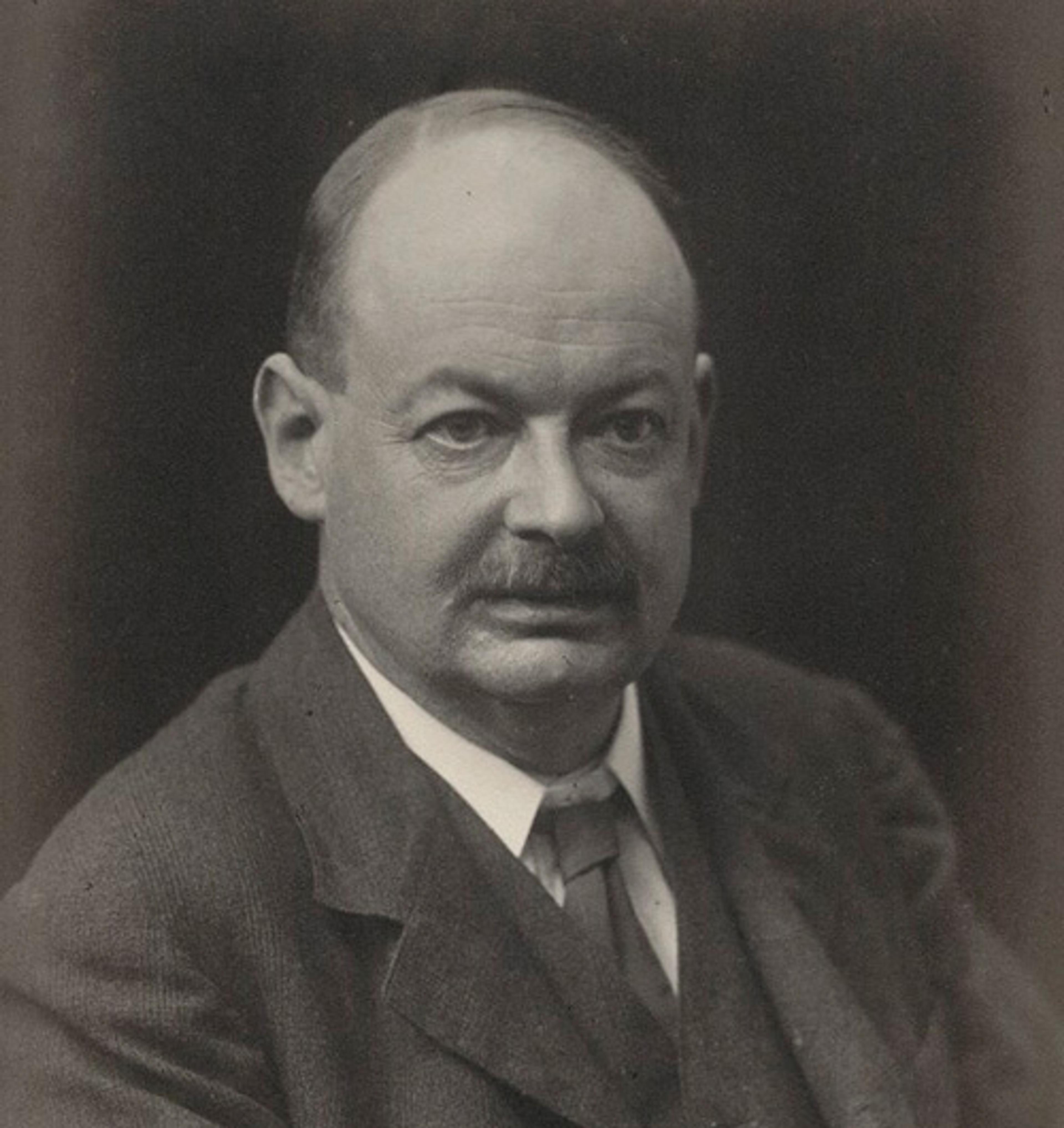 Black-and-white photo of a middle-aged man with a moustache and receding hairline wearing a suit and tie, looking directly at the camera.