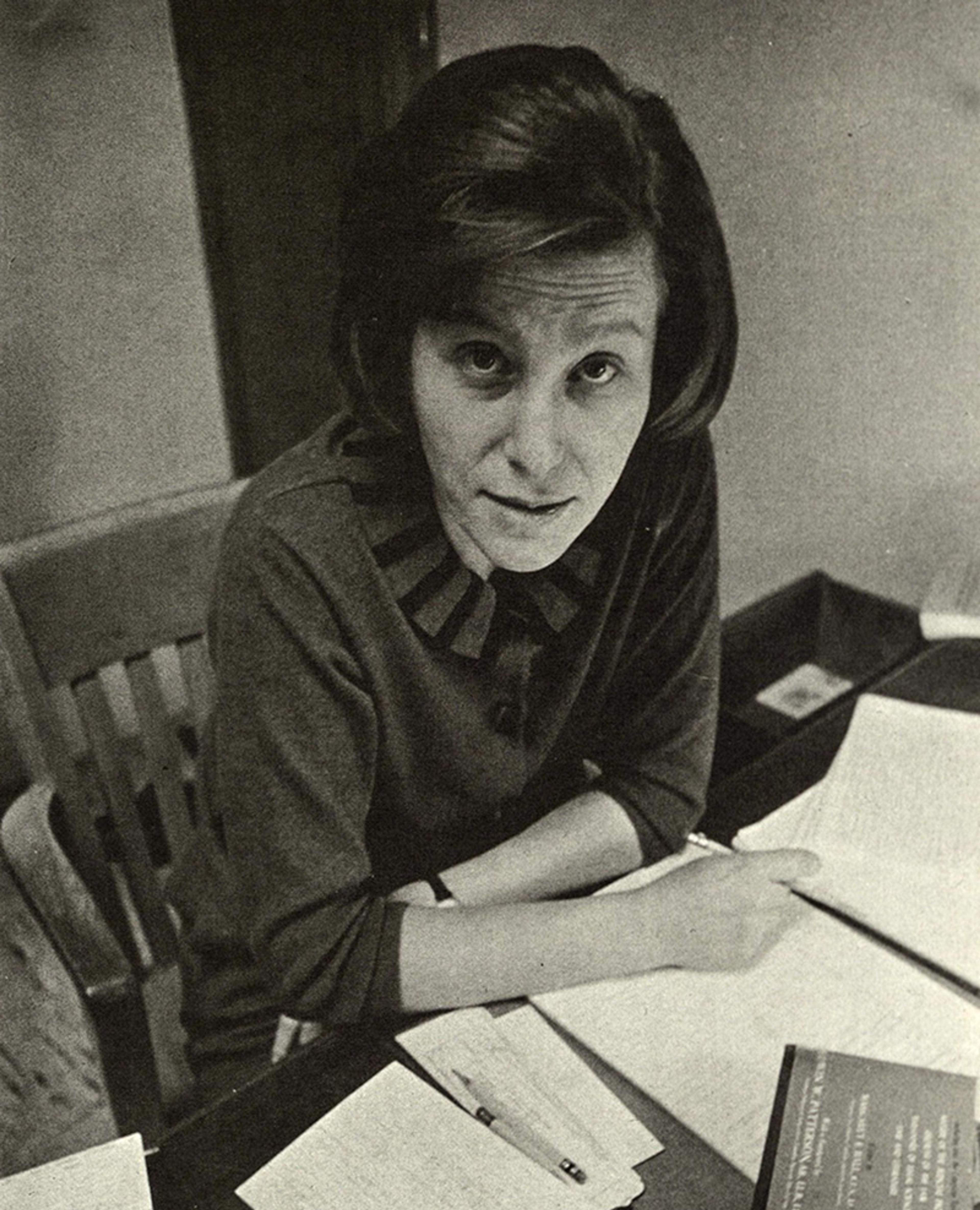 Black-and-white photo of a woman seated at a desk with papers, looking up at the camera, wearing a sweater with a bow design.