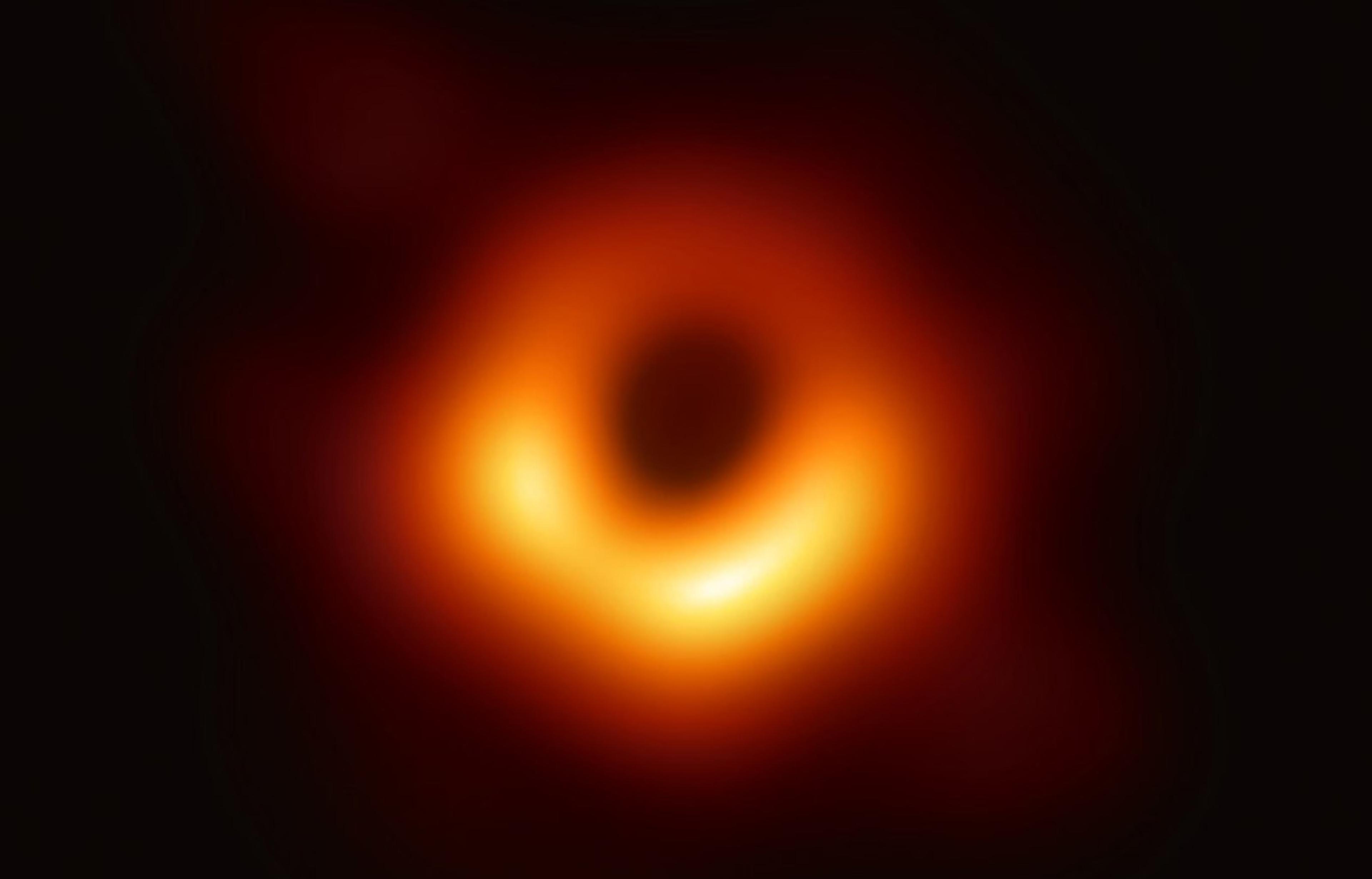 Photo of the first image of a black hole with a dark centre surrounded by a glowing, uneven ring of orange, yellow and red.