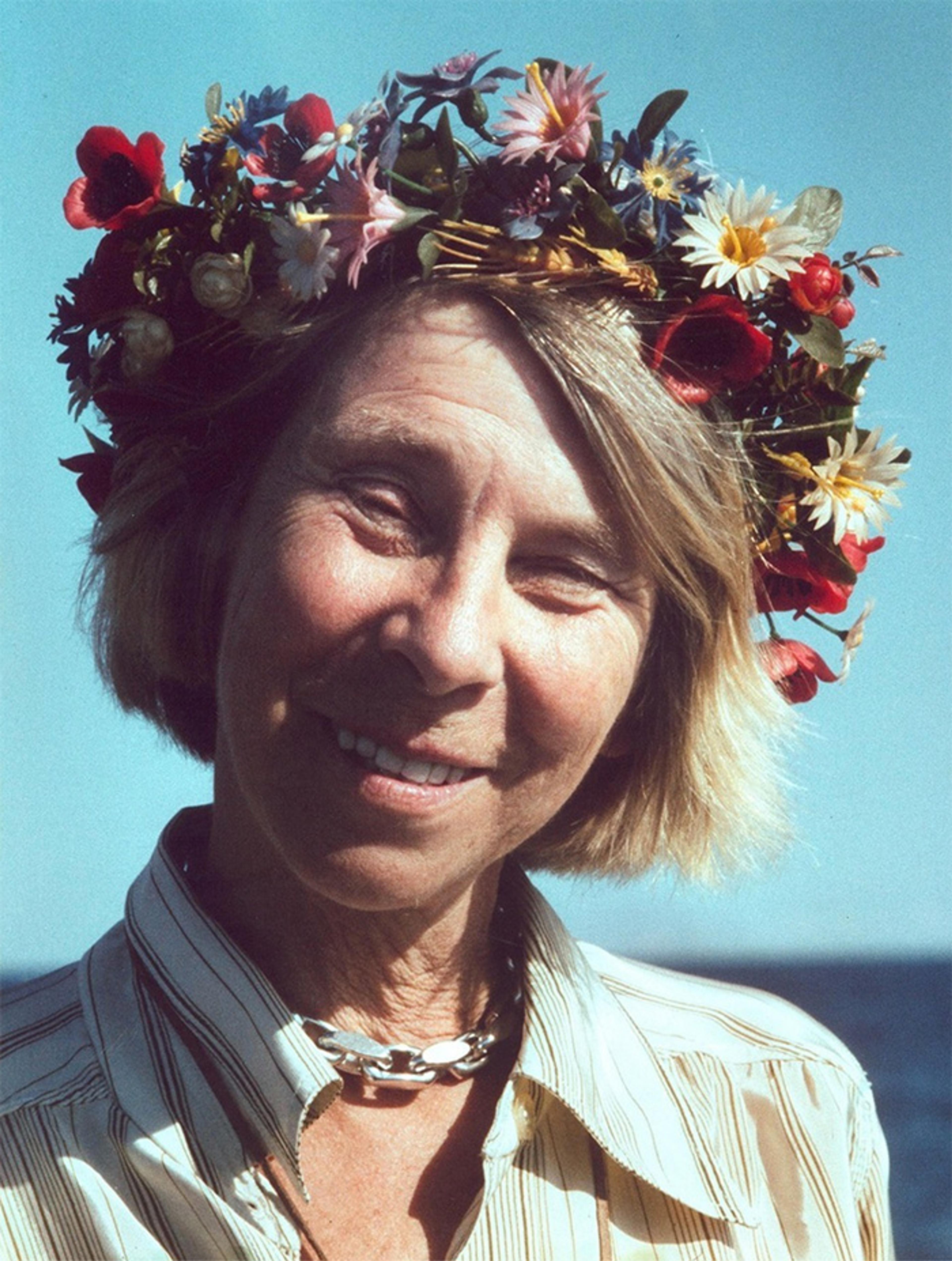 A smiling woman with short blonde hair wearing a floral crown and a striped shirt against a clear blue sky background.