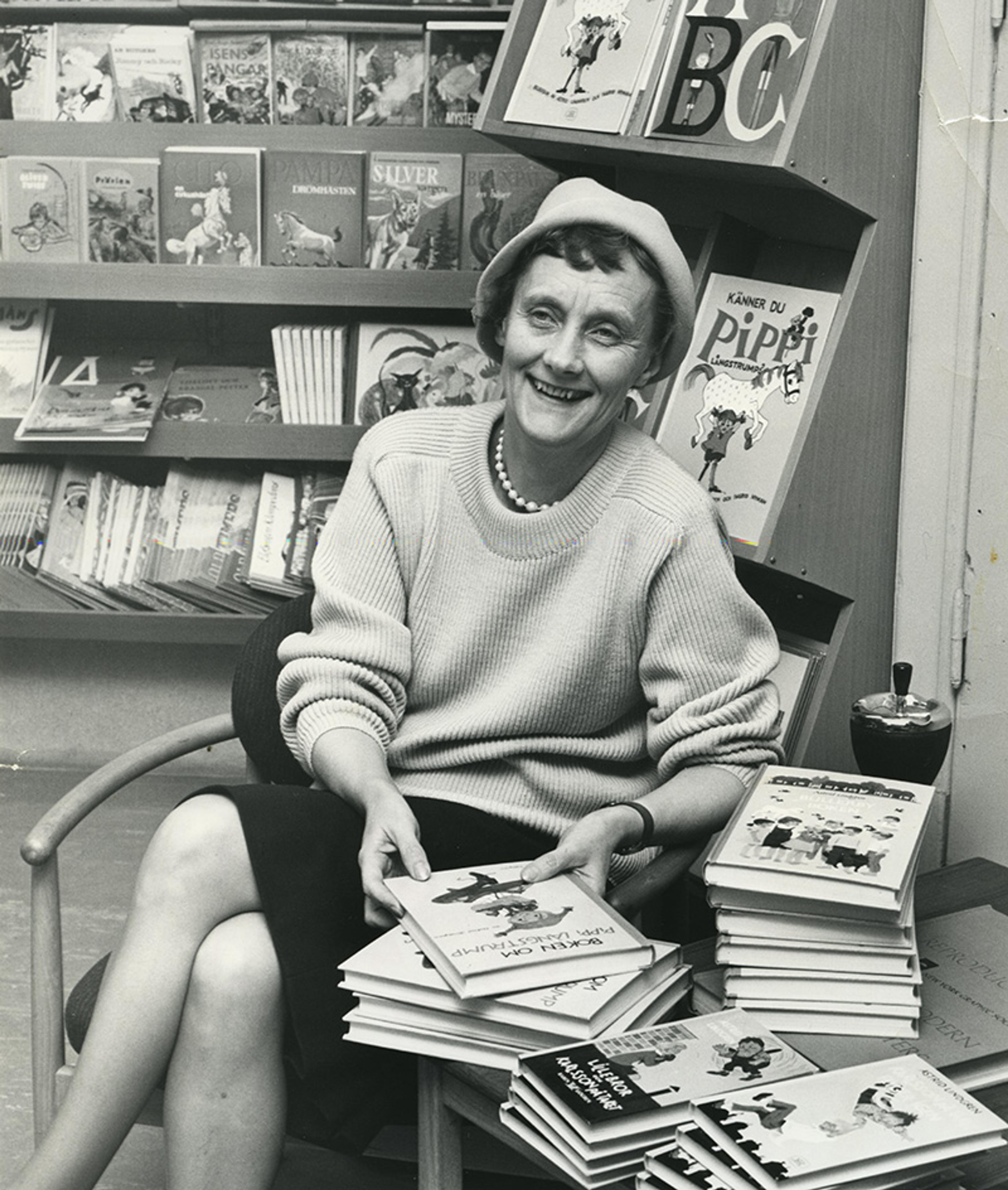 Black-and-white photo of a woman in a hat and sweater, smiling, surrounded by stacks of Pippi books in a bookshop.