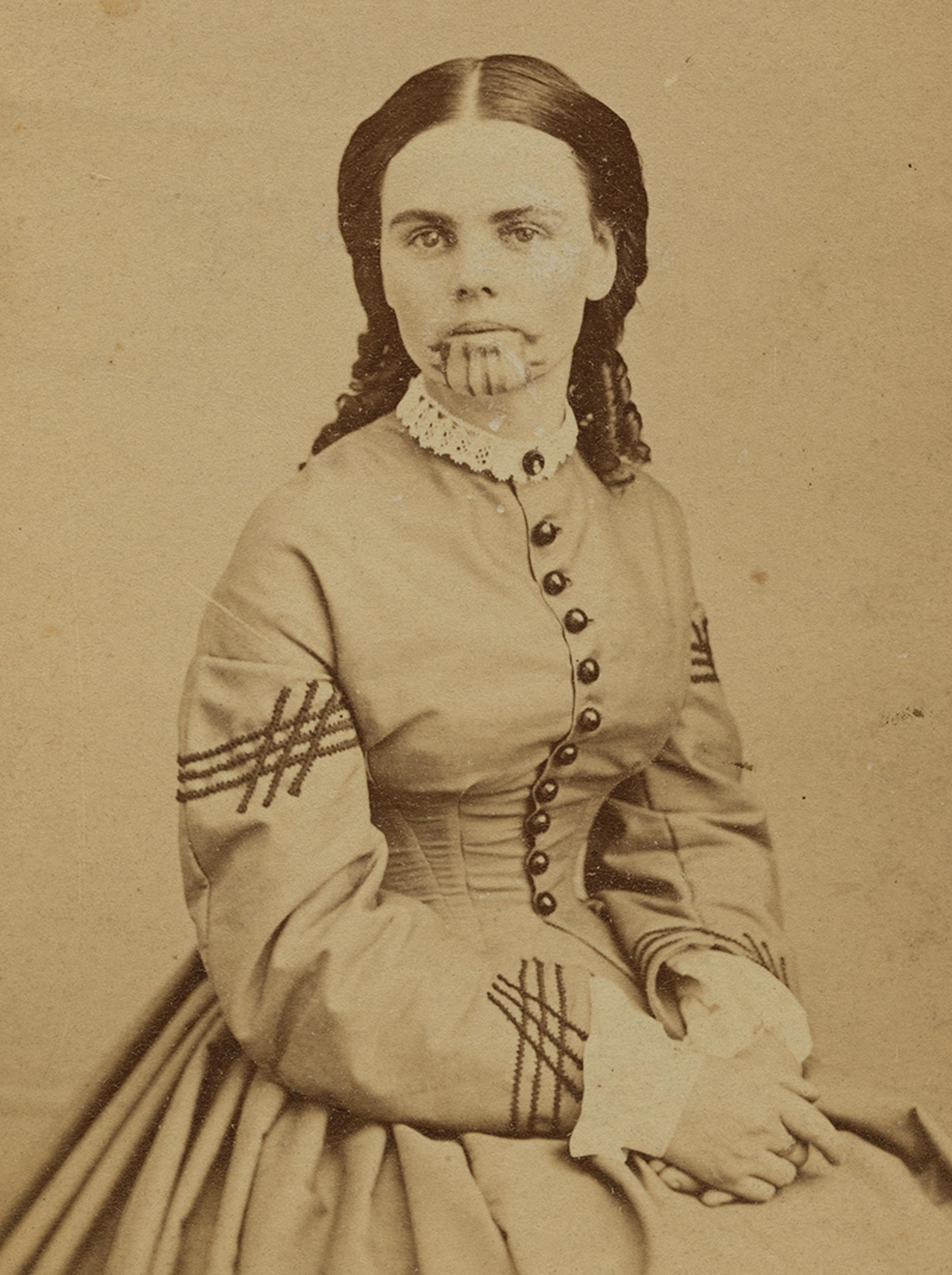 Sepia photograph of a woman in 19th-century attire with traditional chin tattoos; she has her hands clasped in her lap.