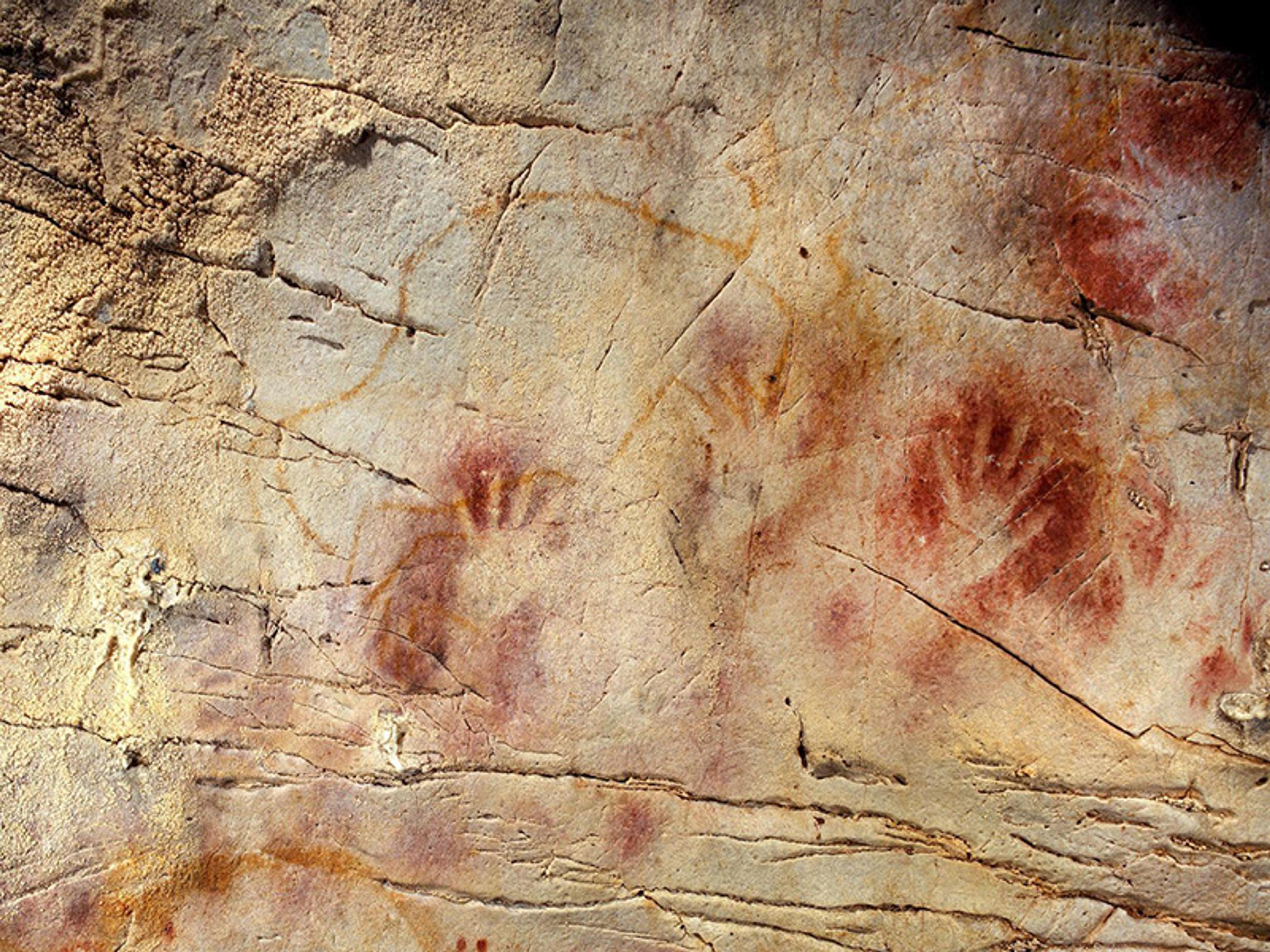 Cave painting with red handprints on a textured rock surface, featuring multiple imprints and faint orange lines in the background.