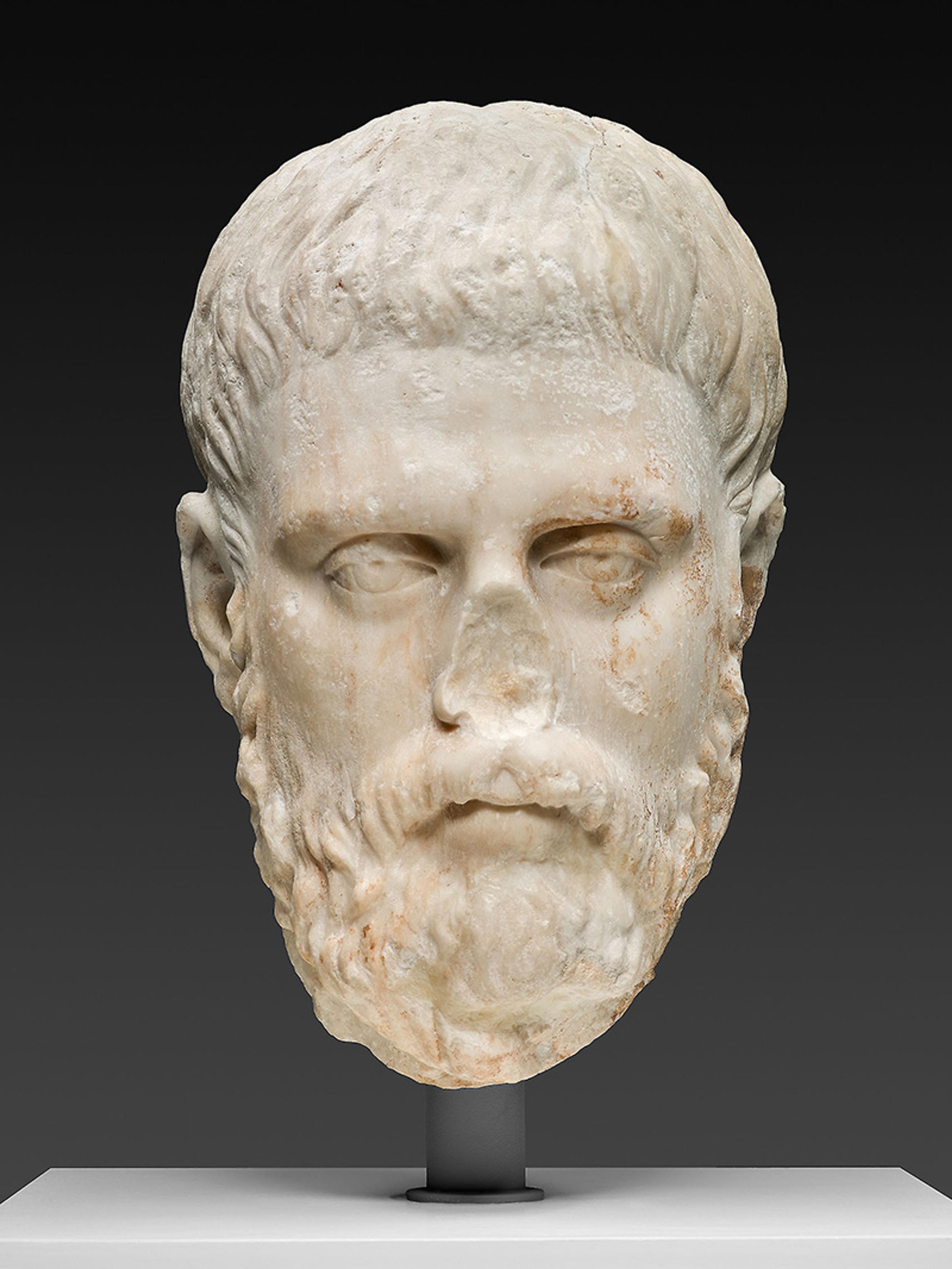 Marble sculpture of a male head with a beard and short hair on a pedestal. The background is dark to highlight the sculpture’s details.