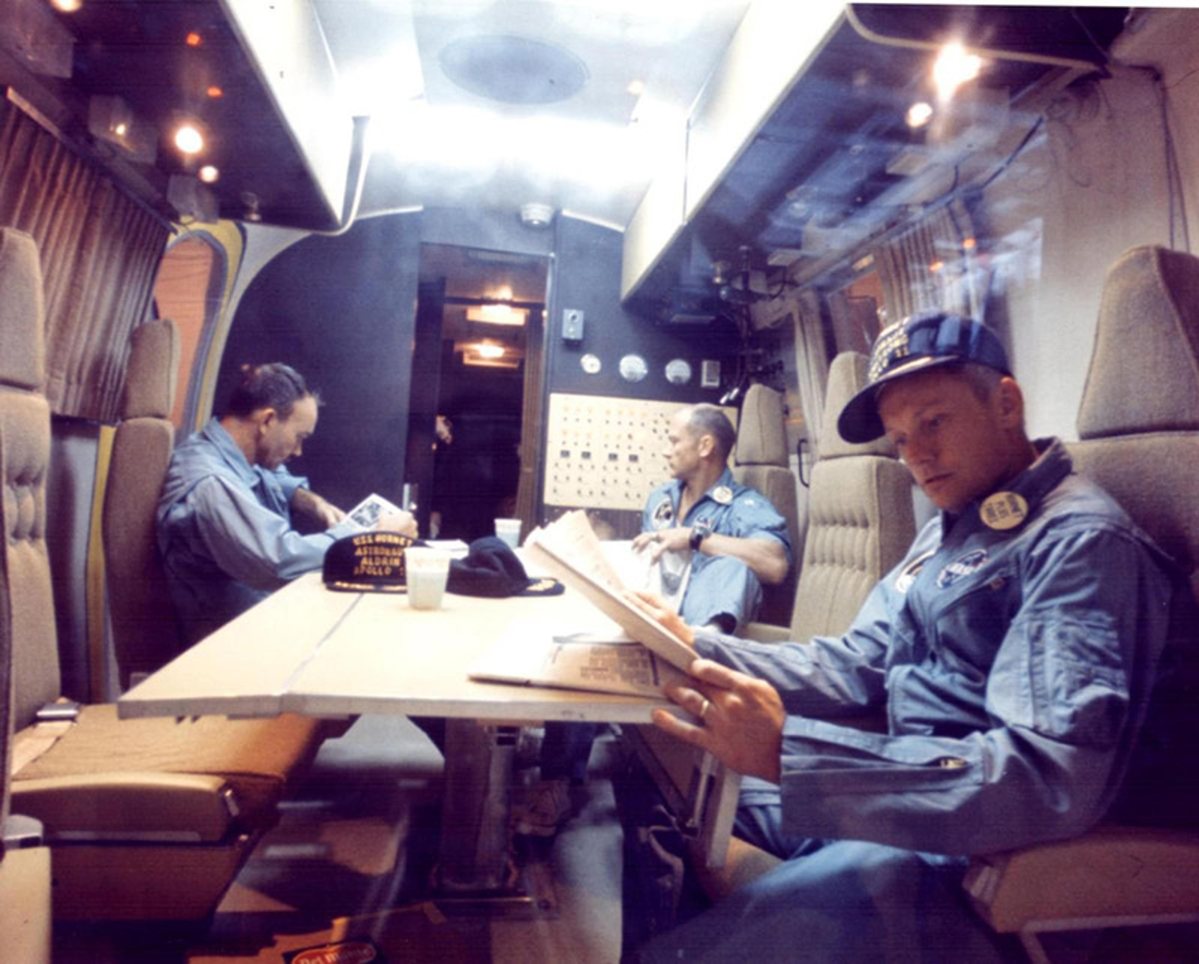 Three astronauts in blue jumpsuits reading inside a cabin with padded seats, a table and a control panel in the background.